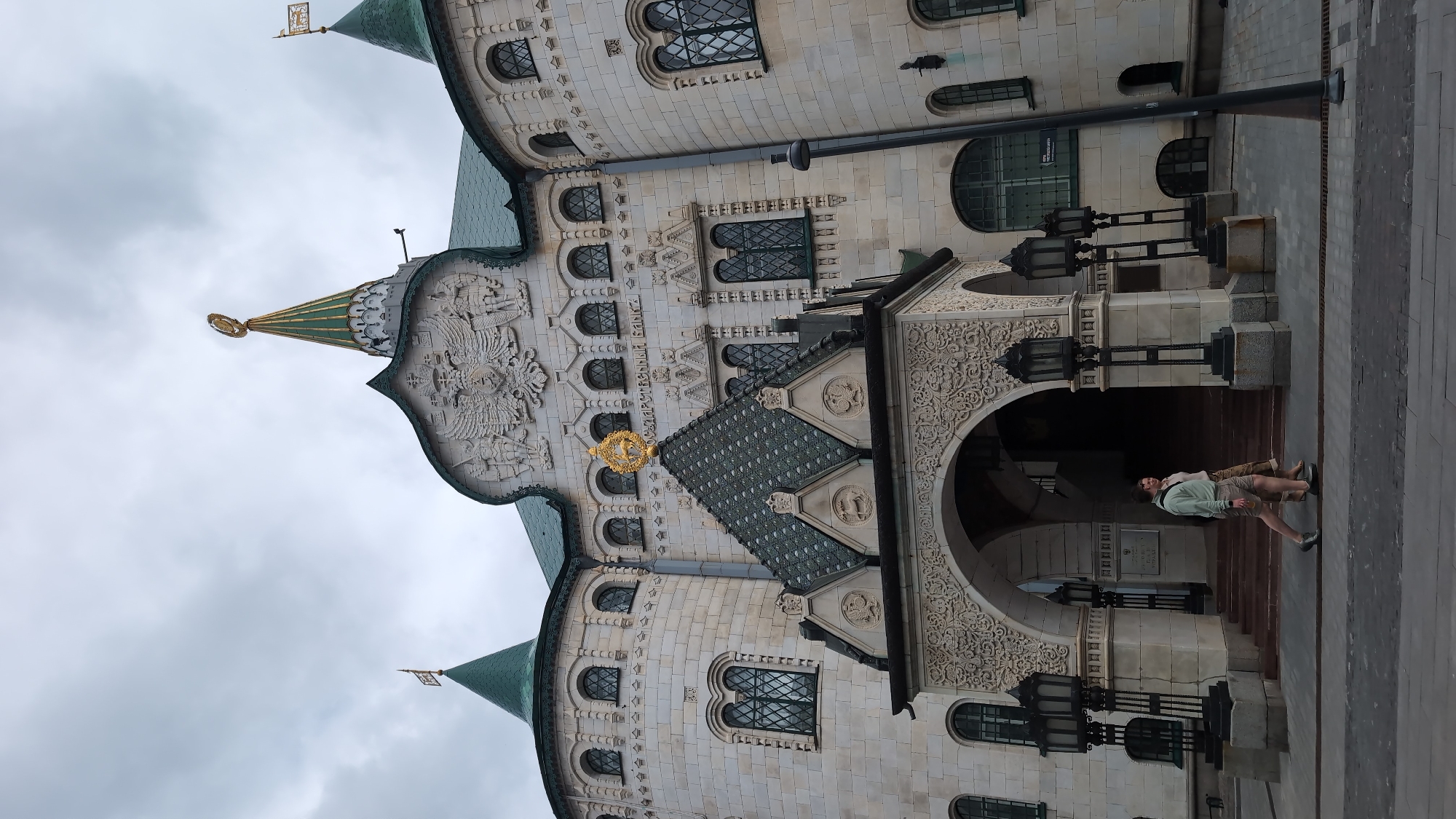 The image captures the Nizhny Novgorod State Bank building, a notable architectural landmark in Nizhny Novgorod, Russia. The building features a distinctive ornate facade constructed from light-colored stone, with a green tiled roof and several towers topped with golden spires. Prominently displayed on the facade is a large relief of a double-headed eagle, a common symbol in Russian heraldry, and the inscription "ГОСУДАРСТВЕННЫЙ БАНК" (State Bank).
In the foreground, two individuals, a man and a woman, are walking across a paved area in front of the building. The man is wearing a light-colored hooded sweatshirt and shorts, while the woman is dressed in a light-colored top and pants. They are both walking towards the left side of the frame. The building is illuminated by several ornate, dark-colored lanterns flanking the entrance. The sky is overcast, suggesting a cloudy day. The overall scene depicts a public space in front of a historically significant building.