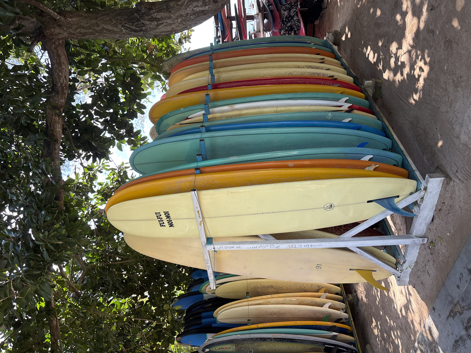 The image shows a tall stack of colorful surfboards vertically arranged on a rack. The surfboards are in various shades, including yellow, orange, blue, and red. The rack appears to be made of metal. Above the surfboards, there is a tree with green leaves.
The image was likely taken outdoors on a sunny day. The location is Kuta Selatan, Indonesia.