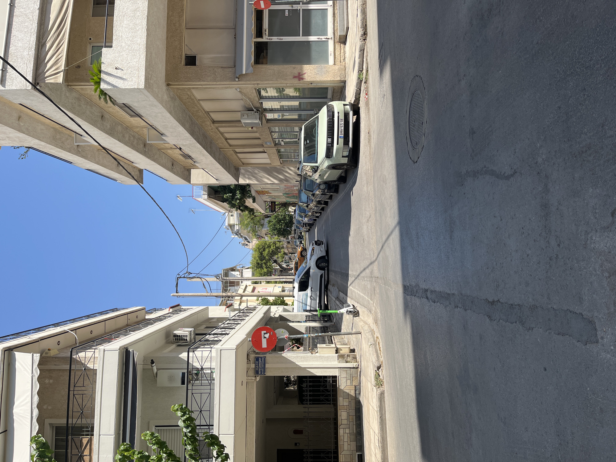 This is a street-level, eye-level, medium shot of a narrow urban street in Athens, Greece. The street is paved, with a visible seam running down the center and a manhole cover in the foreground. Sunlight casts shadows from buildings onto the street.
On the left side of the street, a multi-story building features balconies with black metal railings. A white van is parked on the sidewalk, alongside a green electric scooter. A red circular traffic sign with a white horizontal bar and a diagonal red bar, indicating "No Entry" or similar restriction, is mounted on a pole at the intersection. Above this sign, a street sign in Greek reads "ΟΔΟΣ" (Odos, meaning street) and what appears to be a street name.
On the right side of the street, another multi-story building has a facade with multiple windows and awnings. Several cars are parked in a line along the sidewalk, including a light green Fiat with a license plate BMH 6518. The architecture suggests a mix of residential and commercial spaces. The sky is clear and blue. The scene depicts a typical urban environment with everyday elements of city life.