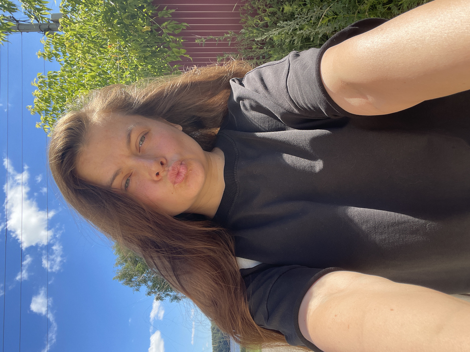 A young woman with long, brown hair poses for a selfie outdoors. She is wearing a black t-shirt and has her lips pursed. The background features a bright blue sky with scattered clouds, green foliage, and a reddish-brown fence. The sunlight suggests it is daytime, likely warm and sunny. The scene appears to be in a rural or suburban area, possibly a backyard or park, given the presence of greenery and fencing. The woman's expression is playful and relaxed, indicating a casual moment captured. There is no visible text in the image.
