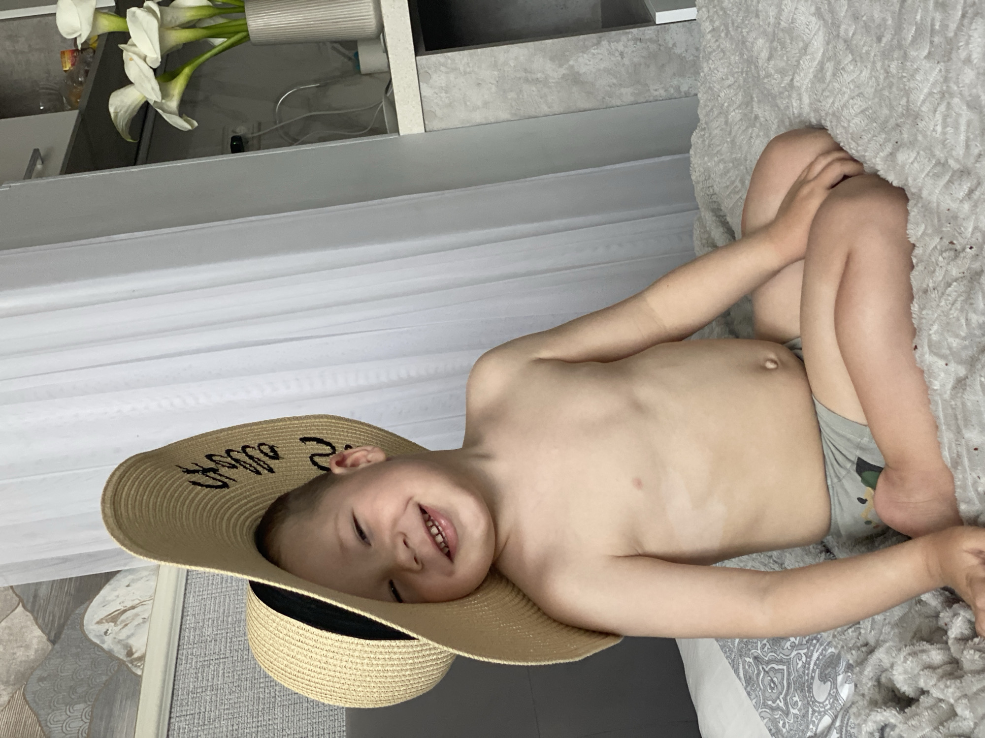 The media file features a young boy lying on a bed. He is wearing a wide-brimmed straw hat with black lettering on the brim that appears to say "Sochi" and a black ribbon around the crown. The boy is smiling and looking towards the camera, appearing happy and relaxed. He is wearing gray shorts with a camouflage pattern. The background shows a textured grey blanket on the bed and a white curtain. In the upper left corner, a vase of white calla lilies is visible. The scene is lit with natural light, suggesting it could be daytime. The presence of the hat and the location cue "Sochi" imply a connection to the Black Sea resort city in Russia, perhaps hinting at a vacation or a warm weather setting.