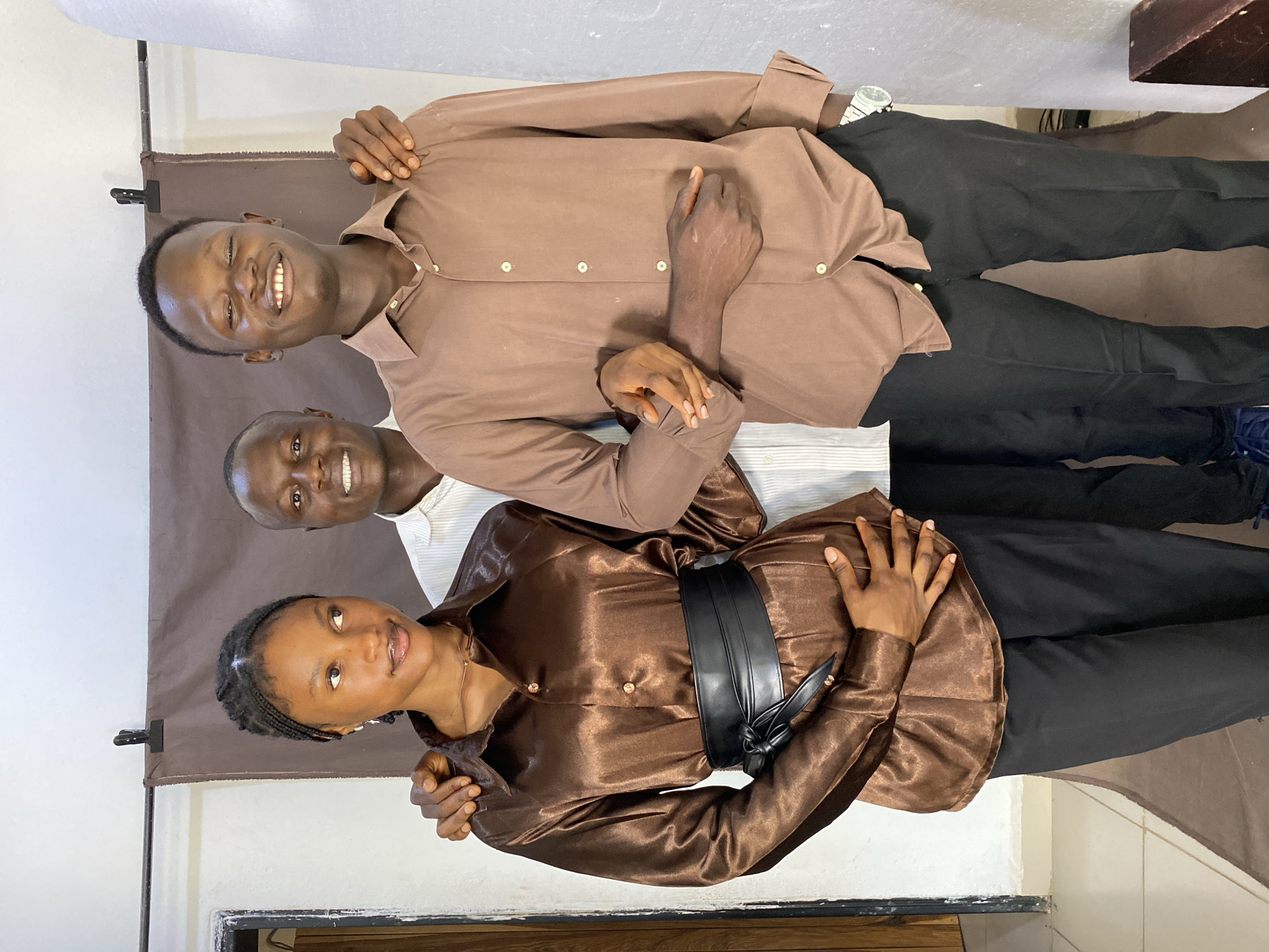 Three individuals, two men and one woman, are posing for a photograph. The woman, on the left, has her hair braided and is wearing a brown, satin-like shirt with a black belt and black pants. She is looking to her left. In the center, a man wearing a white and pinstriped shirt is smiling and looking forward. To the right, another man, wearing a brown shirt and black pants, is also smiling and looking forward. His right arm is around the woman's shoulders, and the man in the center has his arm around the woman's waist. The background consists of a brown cloth backdrop suspended on a horizontal pole, with white walls on either side. The lighting appears to be from an overhead source. The floor visible in the lower left is tiled. The setting is indoors, likely a studio, and is situated in the city of Bukuru, Nigeria.
