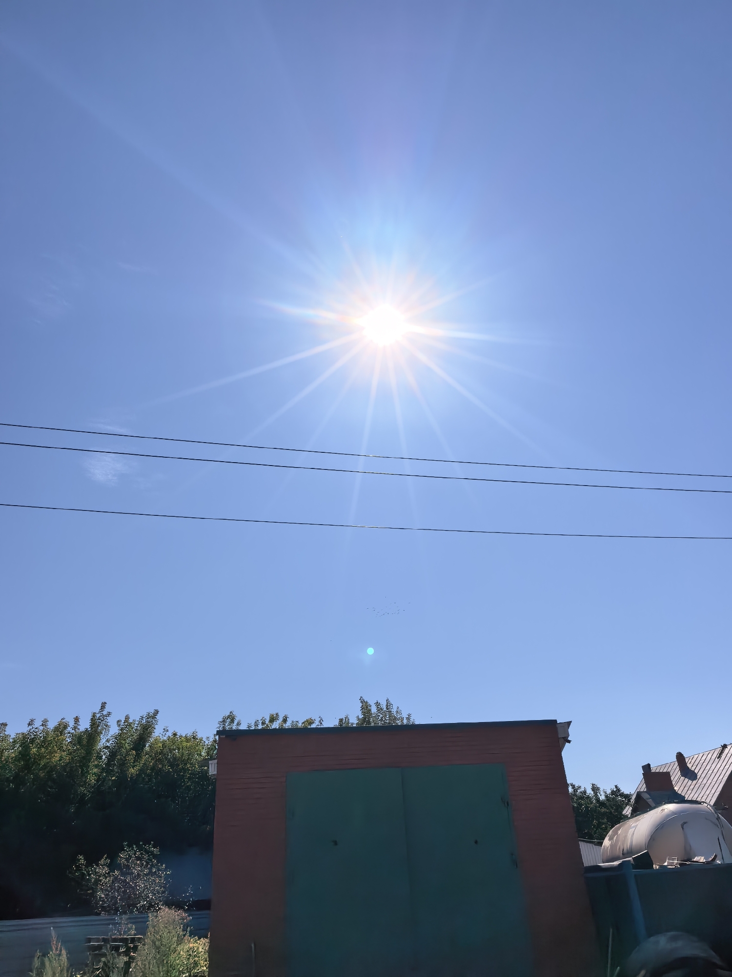 The image depicts a bright, sunny day with the sun prominently displayed in the upper center of the sky, emitting strong rays. Three horizontal power lines stretch across the blue sky. In the lower portion of the image, a brick building with a large, dark green door is visible. To the left of the building, lush green trees and foliage are present. To the right, the roof of a building and a large white cylindrical object, possibly a tank, are visible. A flock of small birds is scattered across the sky. The scene appears to be an outdoor area, potentially in a more rural or suburban setting within the city of Sterlitamak, Russia, given the presence of power lines, a building with a large door, and surrounding vegetation.