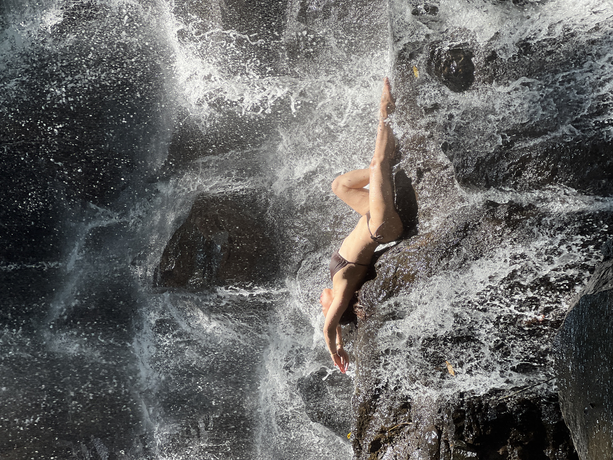 A young woman in a dark brown two-piece bikini is captured in a serene moment, lying on her back on a dark, wet rock surface amidst a powerful waterfall. Her head is tilted back, gazing upwards towards the cascading water, with her arms extended above her head. Her legs are bent, one slightly more extended than the other, creating a relaxed pose.
The scene is dominated by the dynamic movement of water. Turbulent, white foamy water surrounds her, flowing downwards over and around the rugged, dark rocks. Bright sunlight illuminates the waterfall, making the water droplets and spray glisten and shimmer against the dark backdrop of the wet rock face. The contrast between the brilliant white water and the deep, rich tones of the rocks is striking.
The setting is a natural, outdoor environment, typical of a tropical location like Gianyar, Indonesia, suggested by the context. The strong illumination indicates it is daytime. No visible text is present in the image. The overall impression is one of tranquility and connection with nature, perhaps a moment of relaxation or a photoshoot at a scenic waterfall.