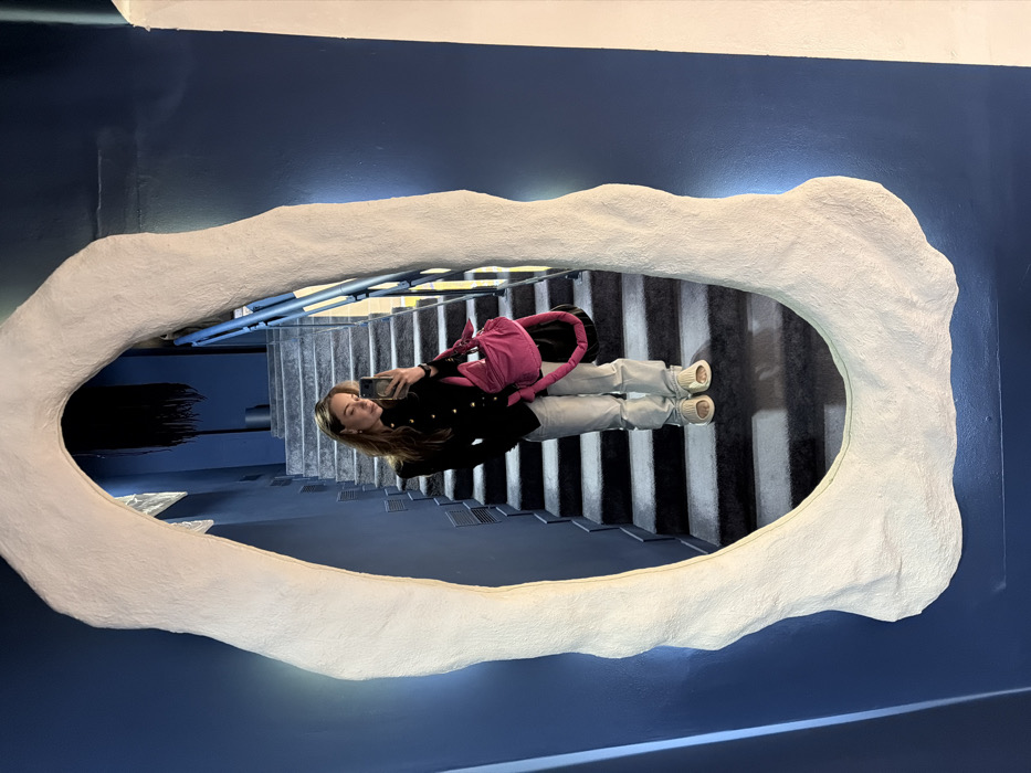 A female subject is depicted in a mirror selfie taken within the Powerhouse Club (Dom Sil Sovetov) in Moscow, Russia. The subject, with medium-length light brown hair, wears a black long-sleeved top with gold buttons, light-wash straight-leg jeans, and cream-colored chunky slides. She carries a fuchsia-colored padded shoulder bag on her left shoulder and holds a smartphone in her right hand, aimed at the mirror. The mirror itself features a thick, irregularly shaped, organic white frame, mounted on a dark blue wall. Reflected in the mirror is a narrow staircase with dark steps and lighter risers, illuminated by ambient light. A blue metal handrail runs alongside the staircase. In the upper background, a dark, textured, possibly textile, art piece is visible. The environment is an indoor space characterized by dark blue walls and muted lighting.