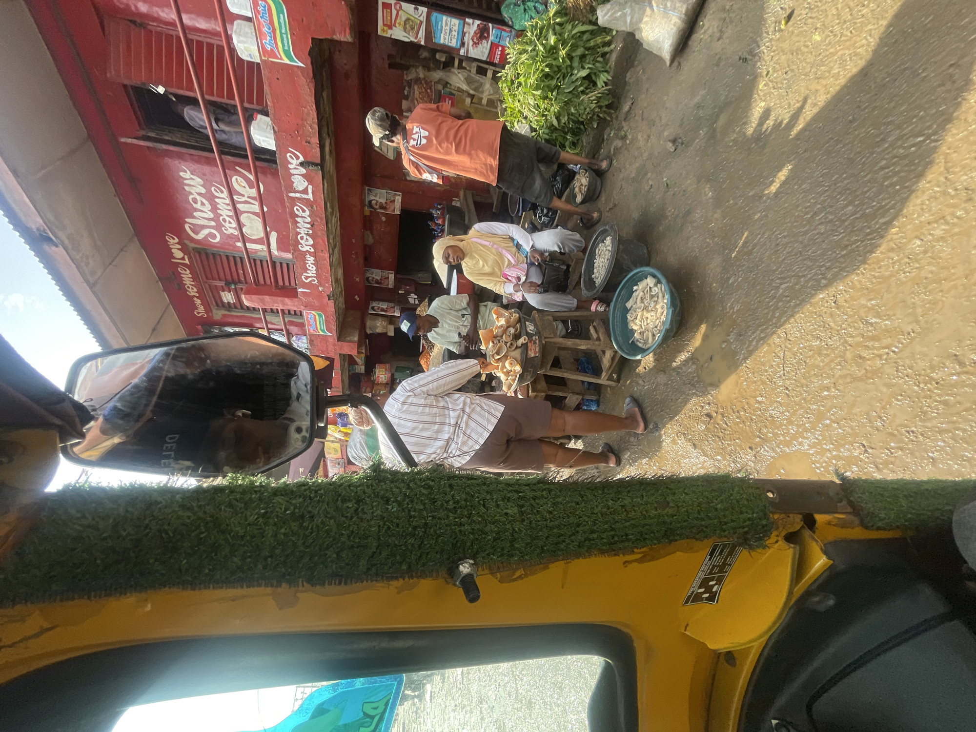 The image depicts a street scene in Akowonjo, Nigeria, likely a market area. A yellow auto-rickshaw occupies the left foreground, its side adorned with artificial grass. In the background, a red building with the words "Show some Love" painted in white is visible. Several individuals are present around a stall. One person, wearing a striped shirt and shorts, stands behind a table displaying food items. Another person, a woman in a yellow hijab, sits near a bucket of food. A man in an orange shirt and cap is seen facing away from the camera. The ground is muddy and wet, suggesting recent rainfall. Various food items and containers are spread out around the stall. The overall impression is a busy, outdoor market environment.
