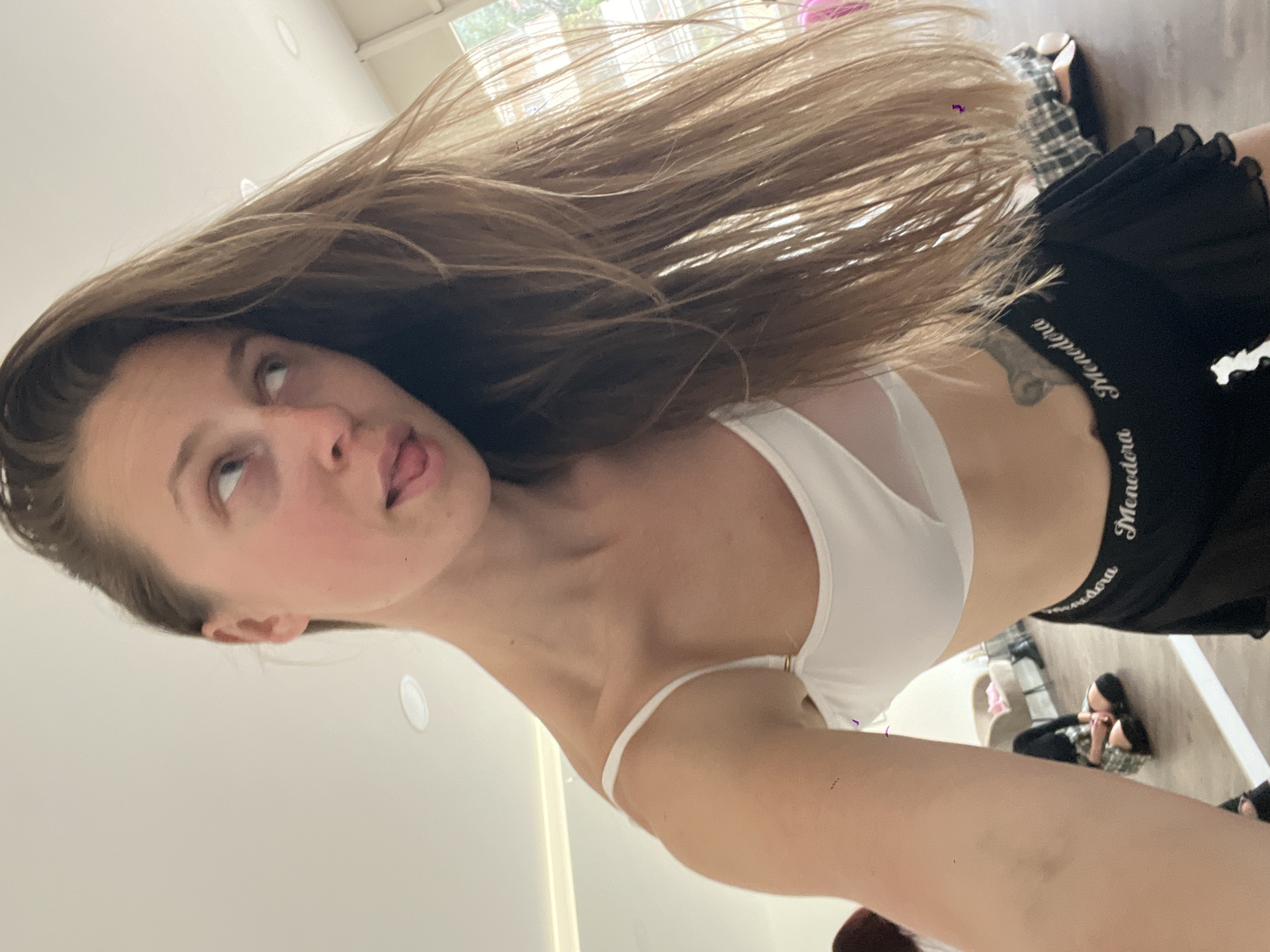 A young female is depicted in a selfie taken from a low angle within an indoor studio. She has long, light-brown hair, worn down, and appears to be in motion as some strands are elevated. Her gaze is directed upwards and to the right, with her tongue slightly extended from her mouth. She wears a white sports bra with thin straps and sheer mesh detailing on the front. Below, she wears a black ruffled skirt or shorts, the waistband of which displays the word "Menodora" in white script repeatedly. A dark tattoo is partially visible on her left side, below her arm. The background features a large mirror reflecting the room, showing a light-colored floor and a white ceiling with recessed lighting. Reflections of other individuals are visible: one person sits on the floor to the left wearing dark attire, and another person sits further back on the right, wearing a plaid skirt. A beige upholstered chair with a pink item on it is also reflected. Through a window or opening on the far right, faint outdoor elements, including greenery, are discernible, indicating daylight. The specific location within Budva, Montenegro, cannot be determined from the visual evidence provided.