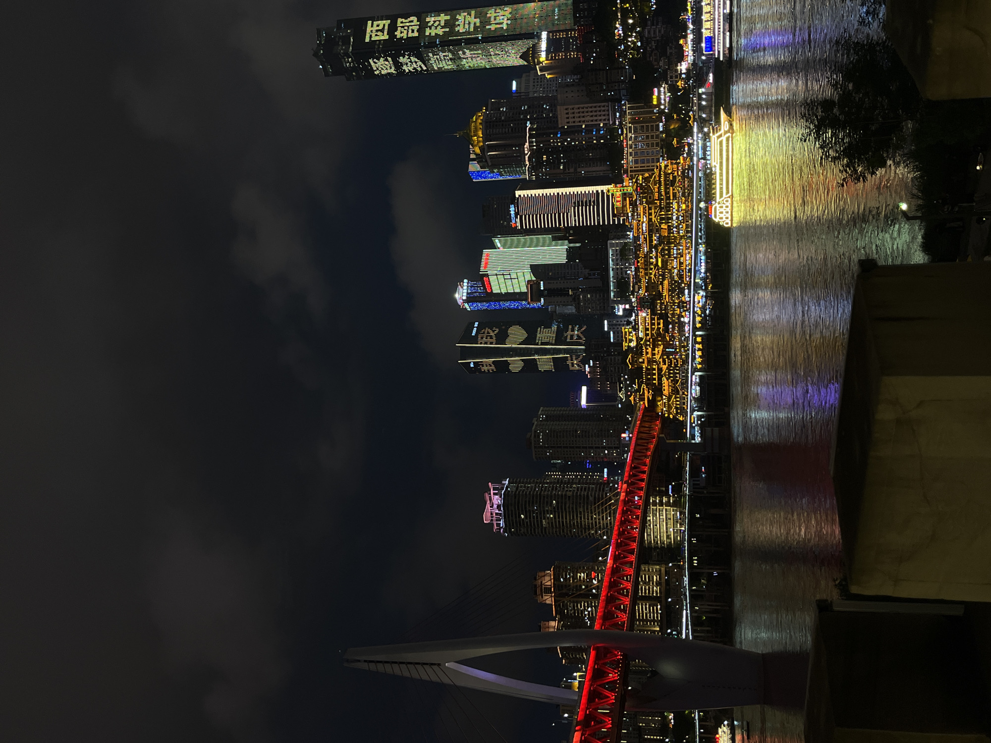 The image shows a night scene of the city of Chongqing, China, specifically the Yuzhong District. A river runs through the middle of the image, with reflections of the city lights shimmering on the water's surface. A modern suspension bridge, illuminated in red, spans the river in the foreground. The city skyline is dominated by numerous high-rise buildings, many of which are brightly lit with colorful lights and displays. The sky is dark, partially obscured by clouds. The composition is vertical.