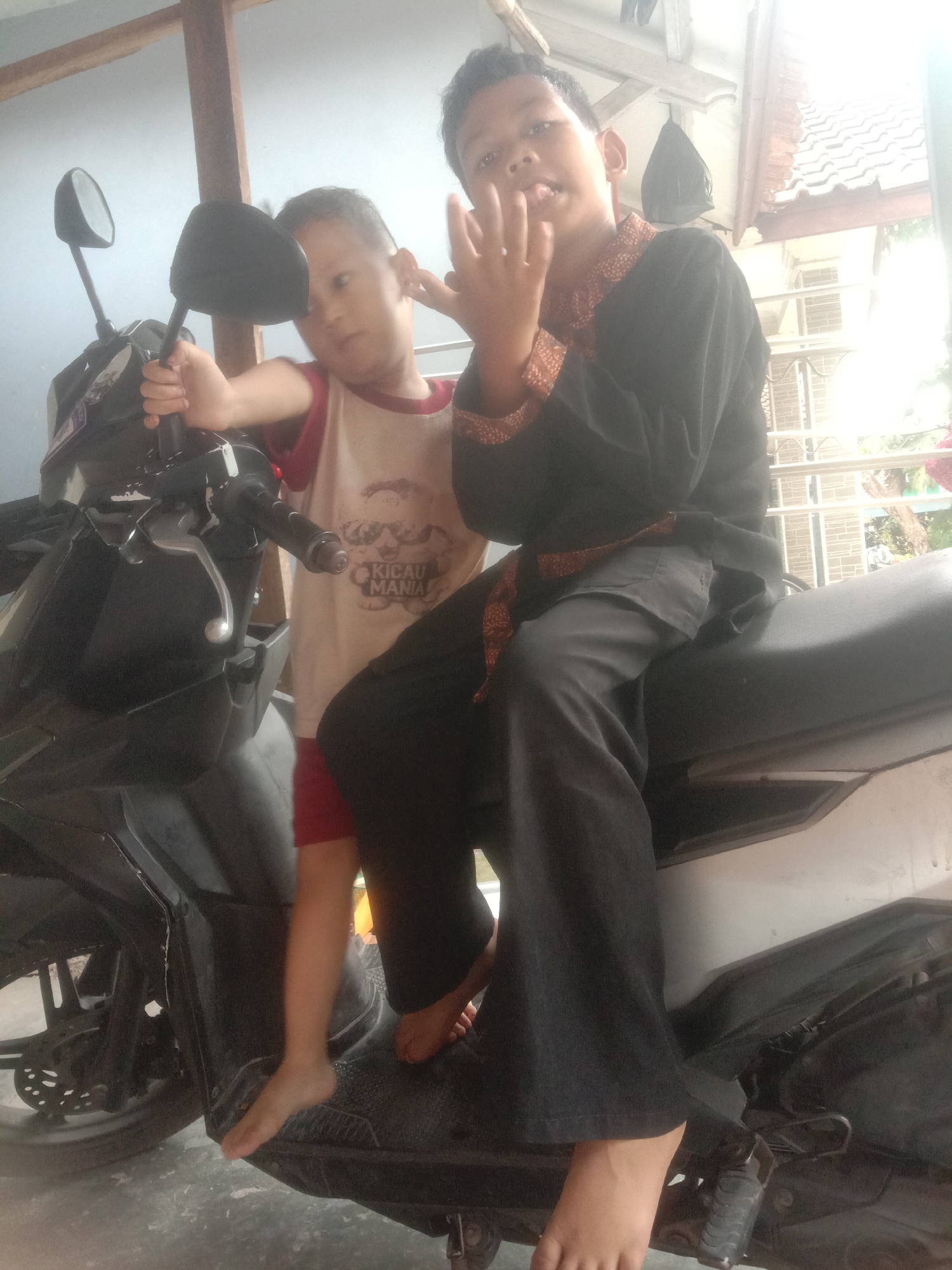 Two young boys are seated on a black motorcycle. The boy on the left is wearing a white t-shirt with red trim and red shorts, and is standing on the motorcycle's footrest. The boy on the right is wearing a black long-sleeved shirt with an ornate red and gold collar and black pants, and is sitting on the seat. Both boys are looking towards the camera. The background shows a simple wooden structure with a tiled roof, consistent with typical housing in Bojong, Indonesia. The lighting suggests it is daytime, likely outdoors or in a semi-open shaded area.