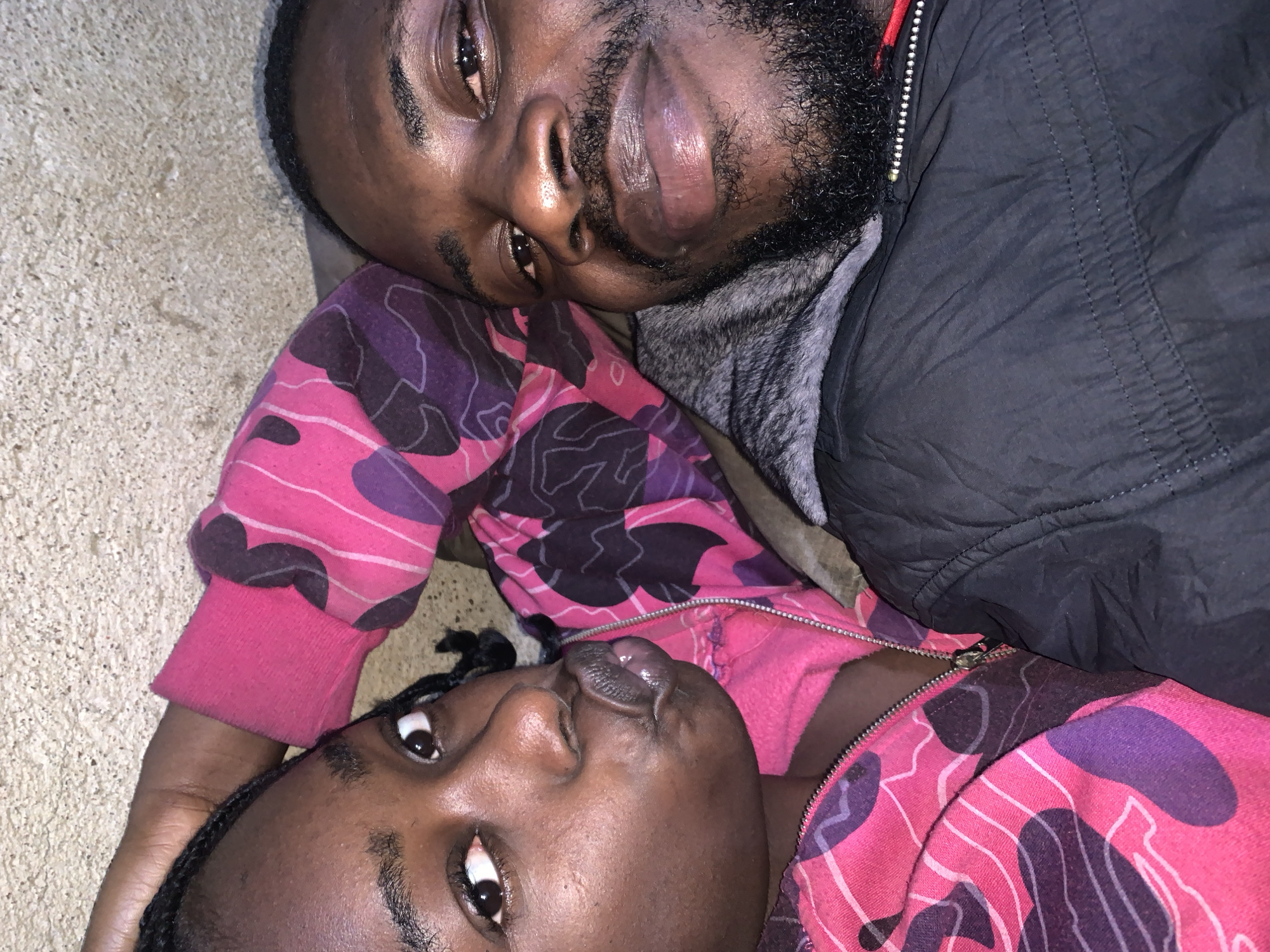 In Rafin Par, Nigeria, a close-up, overhead shot captures two individuals lying down. On the left, a young Black female with braided hair and light brown eyes is looking forward with her lips pursed. She is wearing a pink and purple camouflage patterned hoodie. To her right, a Black male with a full beard and dark brown eyes is looking towards the camera. He is wearing a dark grey hooded jacket with a fur-lined hood. The background appears to be a rough, textured surface, possibly a wall or ground.