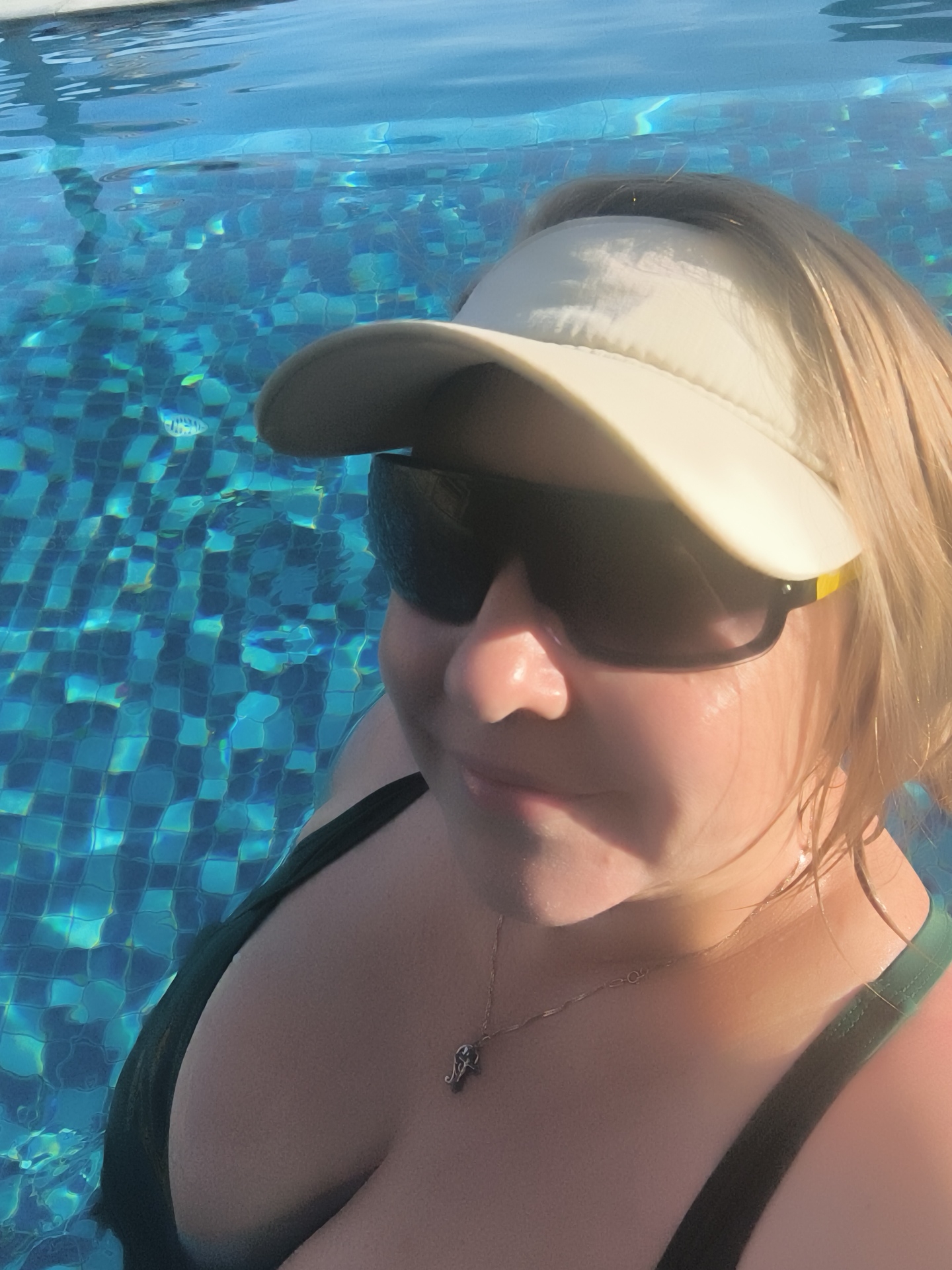 A woman with blonde hair and a tan complexion is in a swimming pool. She is wearing a white visor and sunglasses. She has on a dark green swimsuit and a silver necklace with a small charm. The water is clear blue with a tiled bottom. The lighting suggests it is daytime.