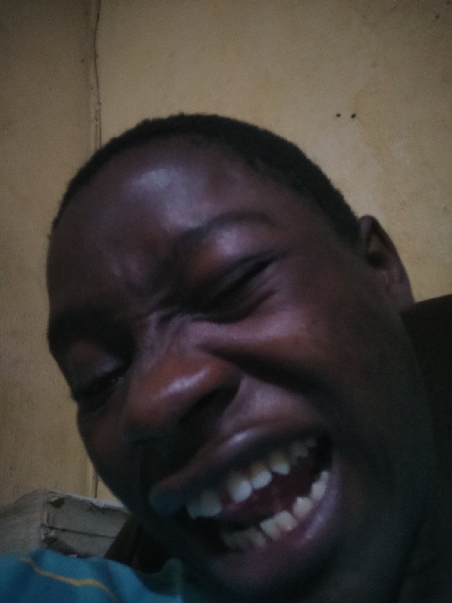 A close-up photo depicts a dark-skinned person, possibly male, with short hair, contorting their face into a wide, exaggerated grin. Their eyes are squinted shut, and wrinkles are visible on their forehead and around their nose. The person's teeth are prominently displayed. The background is a textured, yellow wall. The photo appears to have been taken indoors. There is no information to confirm that the image was taken in Shangisha, Nigeria. The image contains only one visible human subject and is not suggestive of any interactions.