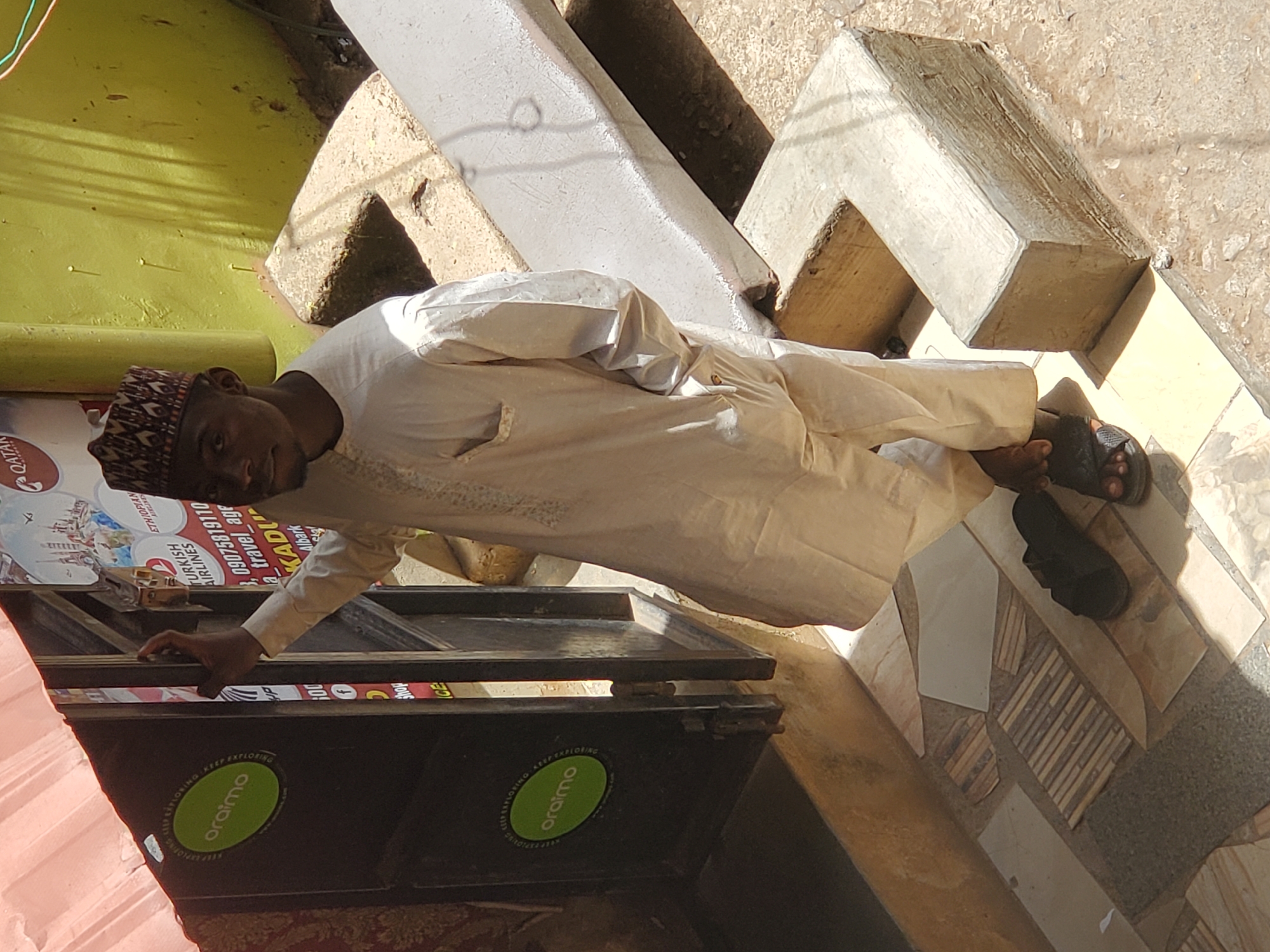 A man is standing next to an advertisement display for oraimo electronics and several airlines, in what appears to be the city of Jos, Nigeria. The man, dressed in a light-colored traditional outfit and a cap, is resting his hand on the display. He is also wearing sandals. Behind him are some concrete structures and the tiled ground. The lighting suggests it's daytime. The advertisement includes airline logos and a phone number starting with "09075819110". A sign is visible with 'Qatar' written on it. A green wall is slightly seen at the back.