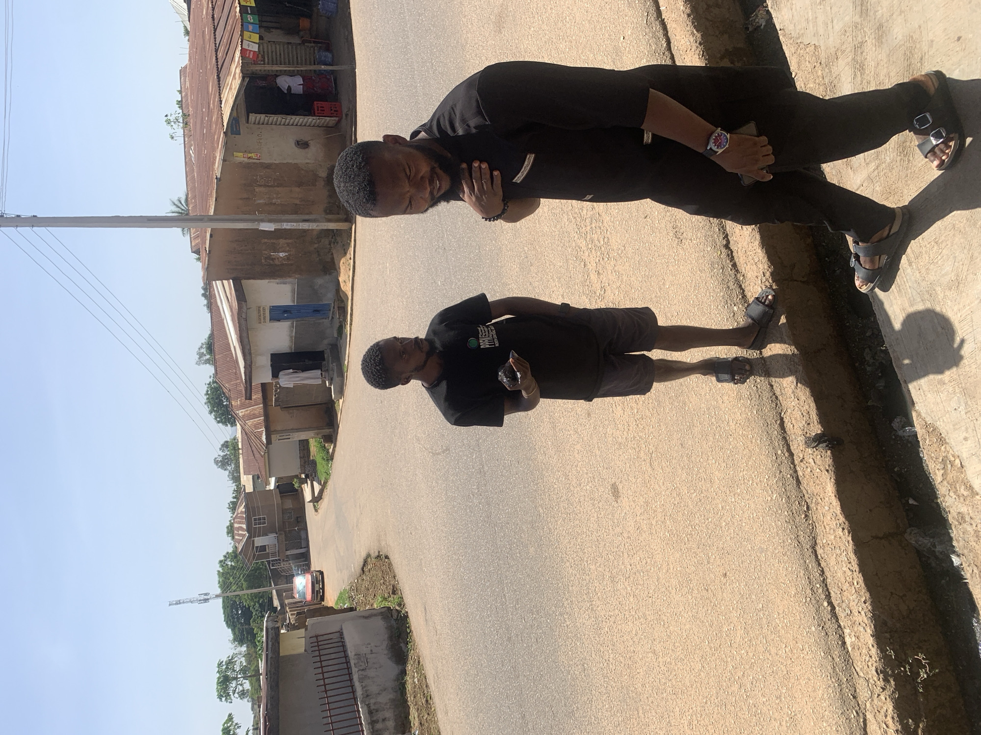 The image captures a sunlit street scene in Jos, Nigeria, featuring two men. The bright, clear sky and sharp shadows suggest strong midday sun.
In the foreground, a man with a beard and short dark hair stands facing the camera. He is dressed in a black long-sleeved shirt, black trousers, and black sandals. His left hand holds a mobile phone, and he wears a watch on his left wrist and a dark bracelet on his right. His right hand rests on his chest.
Behind and to his left, another man, similarly bearded with short dark hair, stands looking towards the camera. He wears a black t-shirt, dark shorts, and black sandals, holding a small dark object in his hands.
The street is an asphalt road with some cracks and an unpaved dirt shoulder. On the left side of the road, several single-story buildings line the street. These structures feature faded walls and corrugated iron roofs, typical of the region. One building has a distinctive blue shutter, and legible text on its facade reads: "RUDLPH ELECTRICAL AND SOLAR COMPANY LTD." Another building appears to be a small shop, with red crates and fabrics visible near its entrance. A tall utility pole with overhead power lines stands prominently beside the road. In the distance, a small red vehicle is partially visible, and sparse vegetation dots the landscape. The overall scene conveys a casual, everyday moment in a local setting.