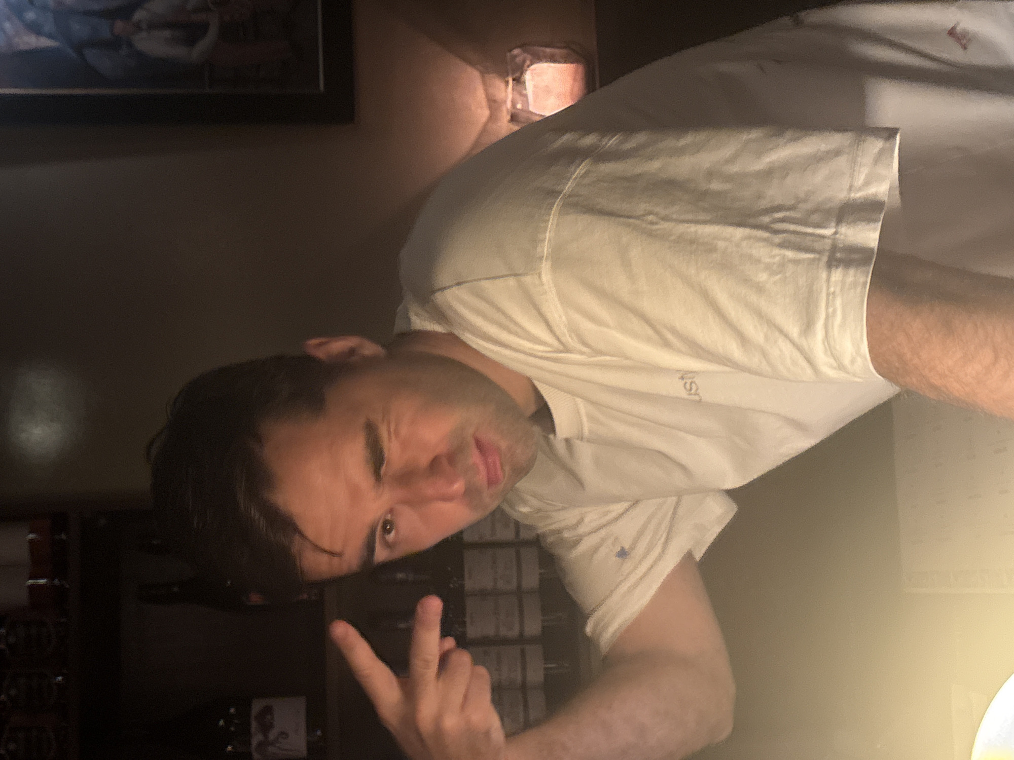A man is posing for a selfie. He is wearing a white t-shirt and making a peace sign with his left hand. He is indoors. There is a dark wooden cabinet behind him with bottles inside. There is a picture on the wall behind him. The lighting in the room is dim. It is impossible to determine the location in Tel-Aviv.