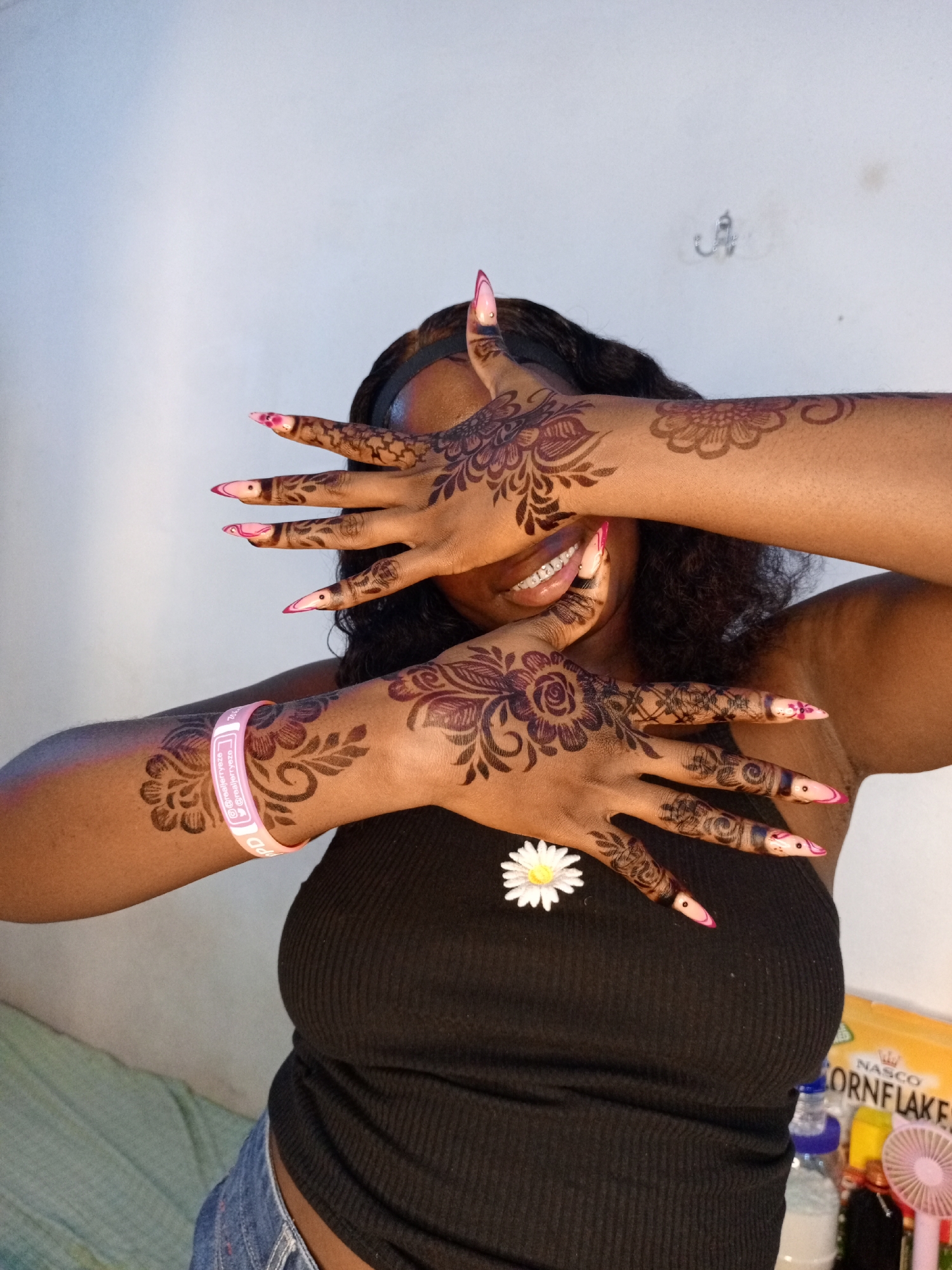 A woman with a black tank top and denim jeans is captured from the chest up. She has dark curly hair and is wearing a black headband. Both of her arms are raised and positioned in front of her face, her hands nearly covering it. Her long nails are decorated with pink and white tips. The woman has dark henna designs on her hands and forearms. A small white flower adorns her tank top. A pink wristband is visible on her left wrist. The background includes a light-colored wall, and slightly out of focus, there are various objects, including a box of cornflakes, a water bottle, and a small pink fan. The location is Unknown, Unknown.