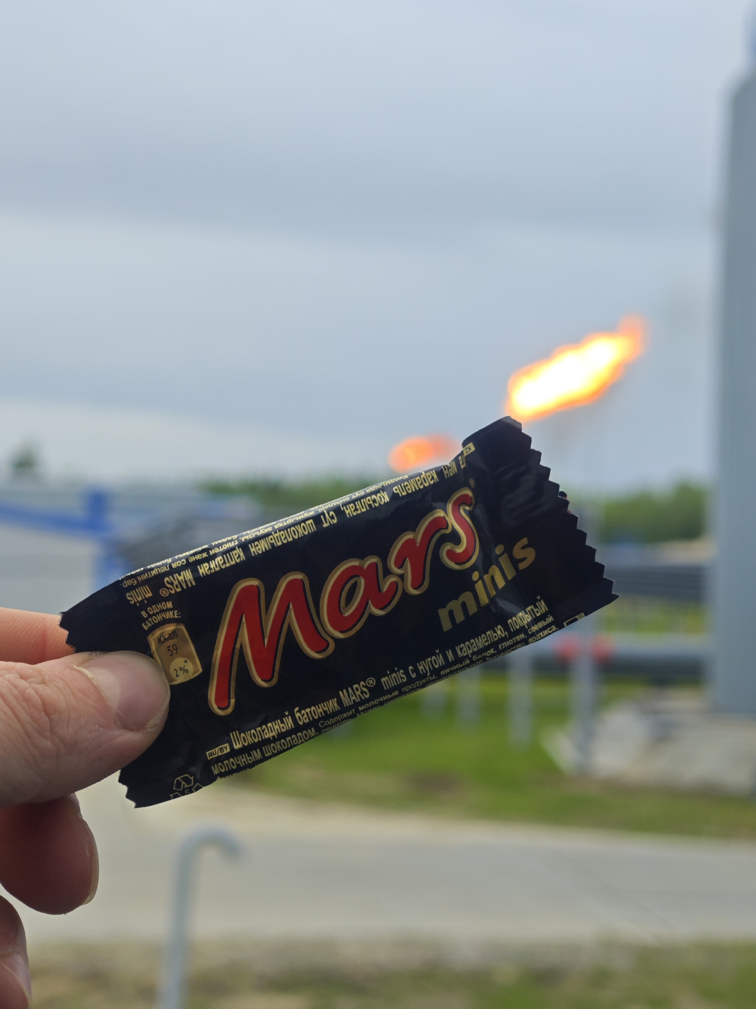 A person's hand holds a "Mars minis" chocolate bar against an outdoor backdrop that includes a blurred flame from an industrial facility. The chocolate bar wrapper is black with the "Mars" logo prominently displayed in red and gold. The wrapper also includes Cyrillic text describing the product. In the blurred background, there is a metal structure, greenery, and a flame appearing to emanate from a stack, suggesting an industrial or manufacturing environment. The sky is overcast, indicating a potentially cloudy day. The setting appears to be on the outskirts of an industrial area.