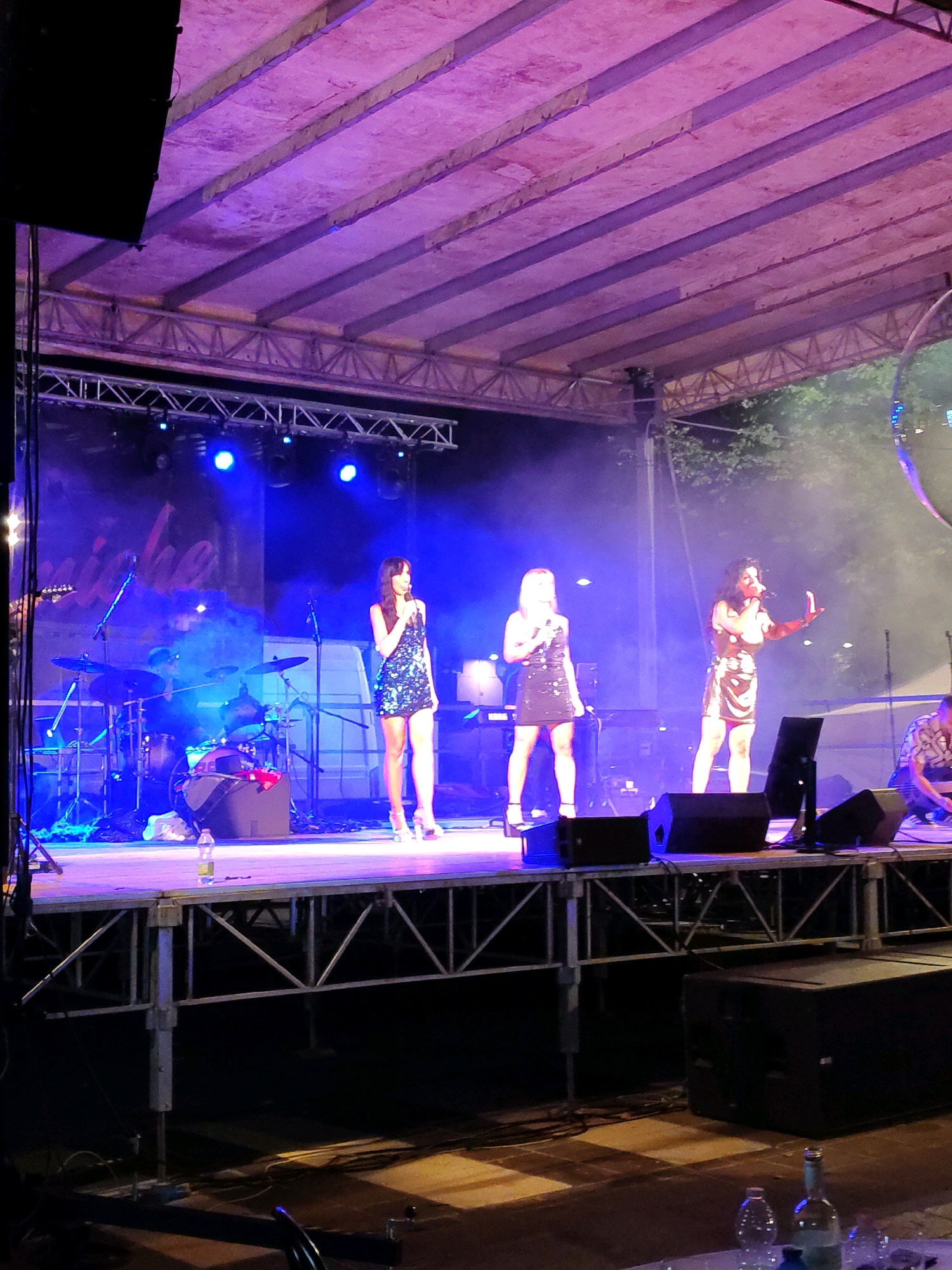 An outdoor concert stage is illuminated by bright blue and purple lights, creating a smoky atmosphere. On the stage, three women are performing. The woman on the far left is holding a microphone and wearing a sparkly black dress. To her right, another woman in a similar sparkly dress is also holding a microphone. The woman on the far right is wearing a gold sequined dress and appears to be gesturing with her hand. A drummer is visible in the background, seated behind a drum set. There are also several musical instruments and audio equipment on the stage. The stage is elevated and appears to be constructed from metal scaffolding. In the foreground, a few water bottles and glasses are placed on a table, suggesting the audience area. The overall setting is a nighttime event with trees visible in the background.