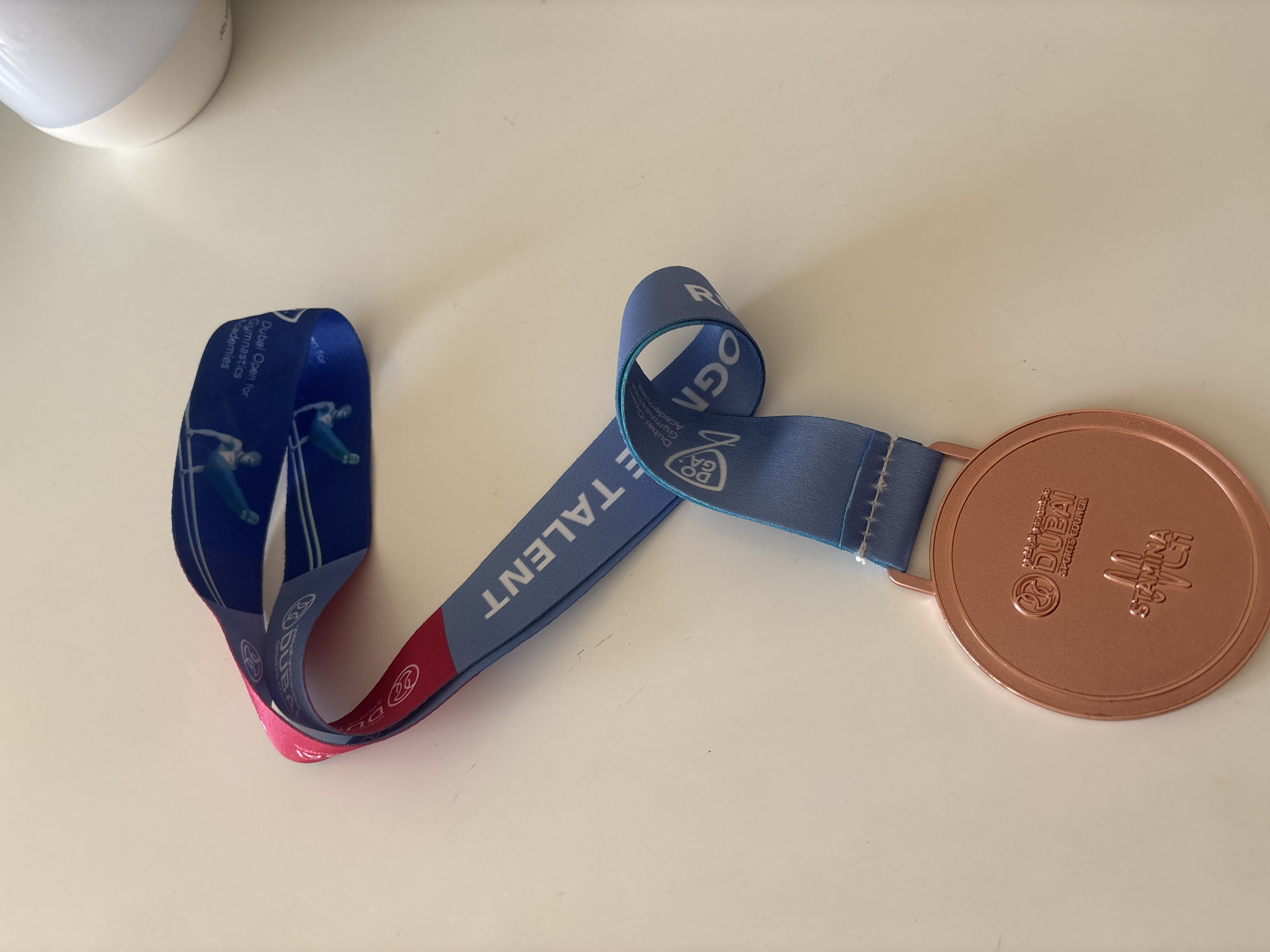The image displays a bronze medal with a blue and pink ribbon, lying on a white, textured surface, likely a tabletop. The medal has embossed text and logos, including "DUBAI SPORTS COUNCIL," and what appears to be a stylized representation of a gymnast. The ribbon is adorned with text and graphics related to gymnastics, specifically mentioning "Dubai Open for Gymnastics Academies" and "TALENT." The lighting suggests an indoor environment. The overall scene is static, with no people or active events depicted.