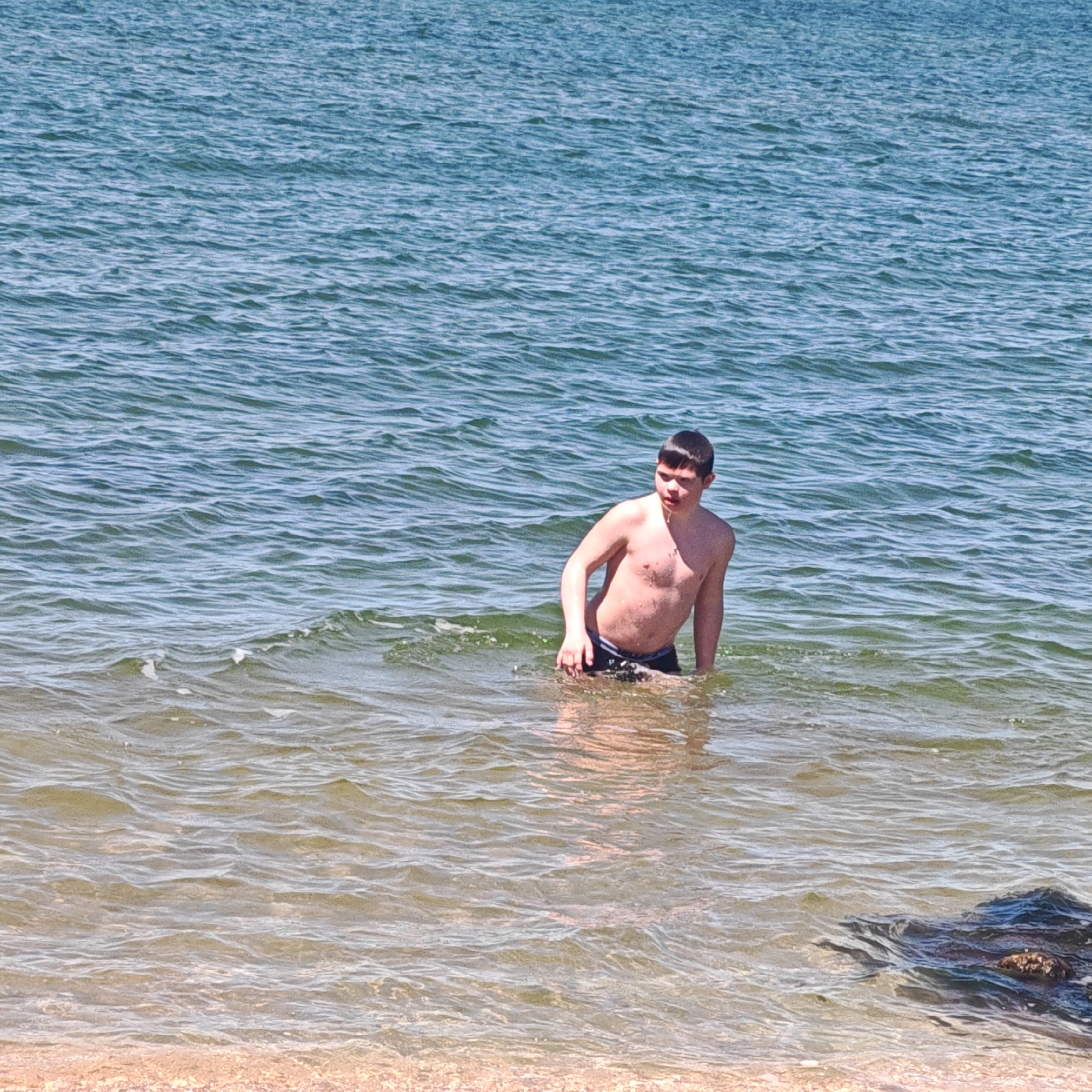 A young, fair-skinned boy stands in the water at a beach in городское поселение Щелкино, Ukraine. He is bare-chested and wearing dark-colored swim trunks. The water reaches about mid-thigh. The sea behind him is calm and slightly choppy. Some rocks are visible in the lower right corner, likely near the shoreline. The sand of the beach is light-colored and appears wet. The scene is lit by bright sunlight.