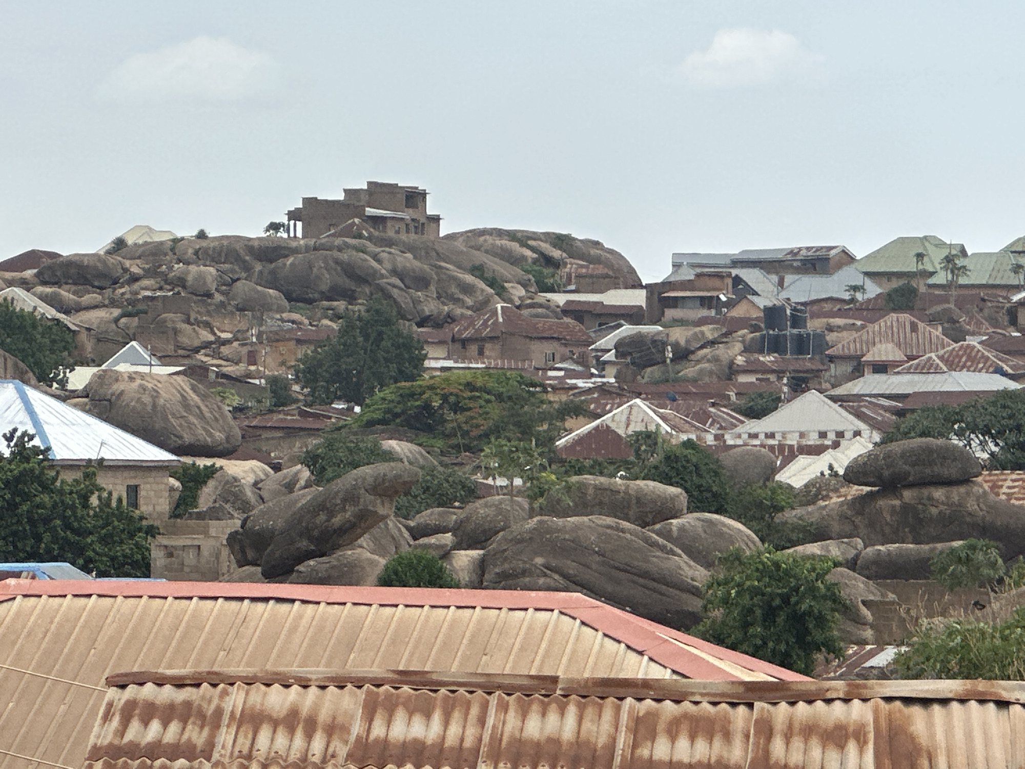 The image depicts a dense settlement in Jos, Nigeria, characterized by structures built amidst and upon large, smooth, gray boulders. Housing varies, with roofs constructed from corrugated metal sheets, exhibiting shades of red, brown, and blue. A multi-story building sits atop the rock formations, blending with the natural landscape. Trees and other vegetation are interspersed throughout the settlement. The sky is overcast, with a pale hue. The foreground features a rusted corrugated metal roof.