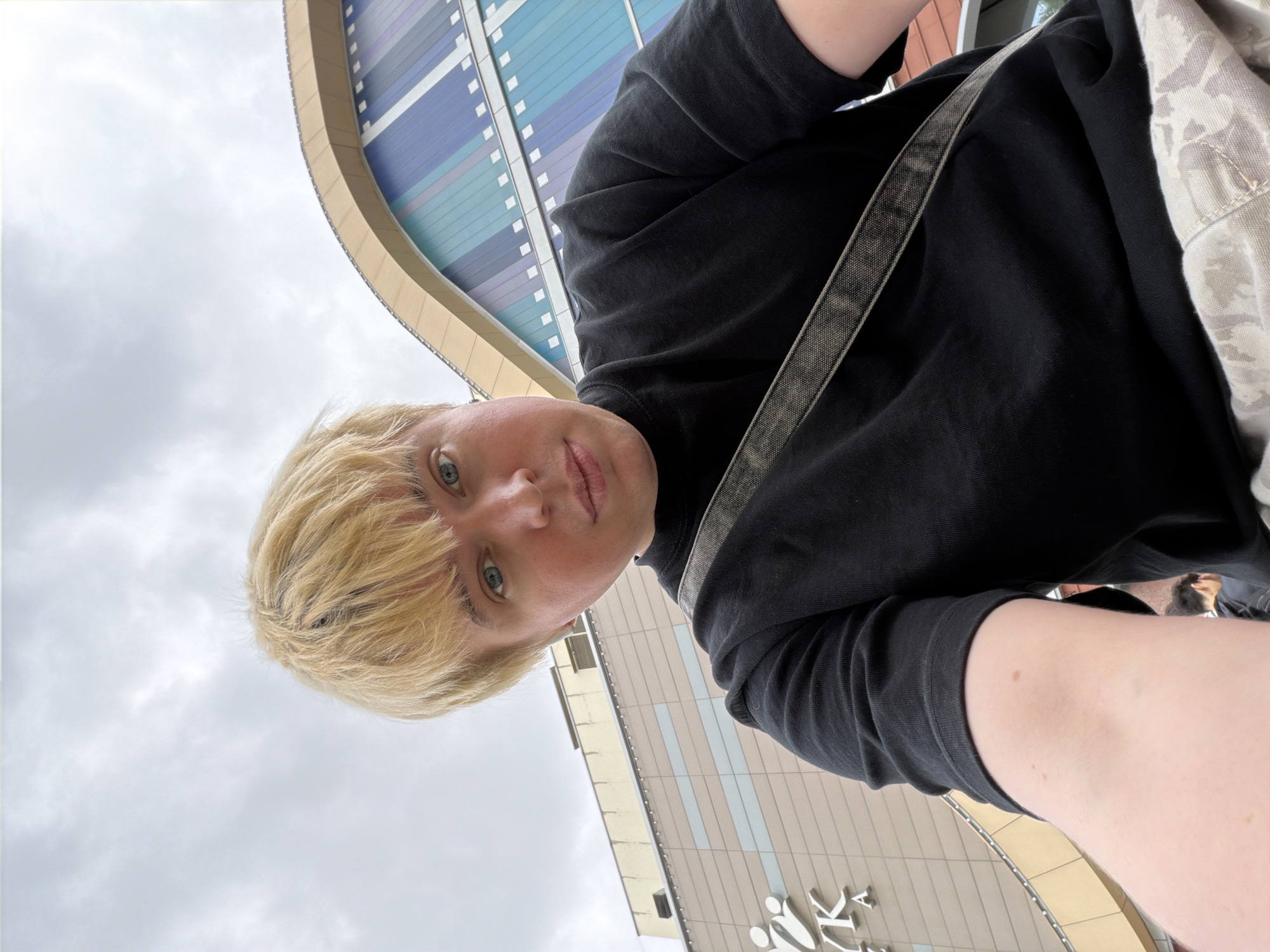 A low-angle shot features a person with short, blond hair and blue eyes, positioned against an urban backdrop. The person is wearing a black top, and a strap of a bag is visible diagonally across their chest. The background showcases modern architecture with colorful panels and an overcast sky. A building with a recognizable symbol or logo on its lower portion is also visible. The location appears to be an outdoor public space in a city, Unknown, Unknown.