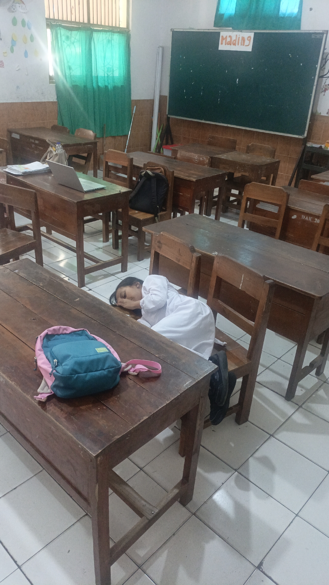 The image depicts a classroom setting in Salatiga, Indonesia, with a young girl wearing a white uniform lying asleep on a wooden desk. Her head is resting on her arms, and her face is partially obscured by her hair. A teal and pink backpack is placed on the desk in front of her. The classroom is furnished with multiple wooden desks and chairs, some of which appear old and worn. In the background, a green chalkboard with the word "Mading" written on it in orange is visible. Sunlight streams through a window covered by green curtains, illuminating the room. The floor is tiled with white squares. The overall impression is one of a typical, perhaps slightly dated, Indonesian schoolroom, where a student has succumbed to fatigue during the school day.