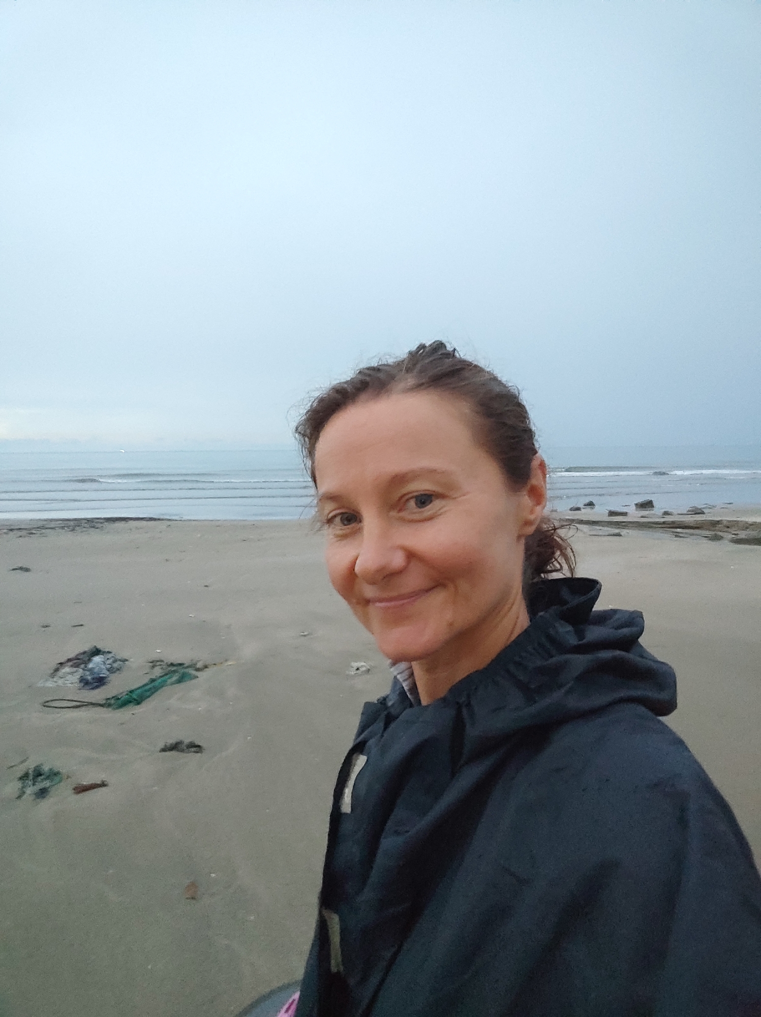 A woman is standing on a sandy beach. The ocean is in the background with waves gently rolling in. The sky is overcast and gray. There is some debris on the beach. The location appears to be a coastal area, with no specific information available to pinpoint the city and state.