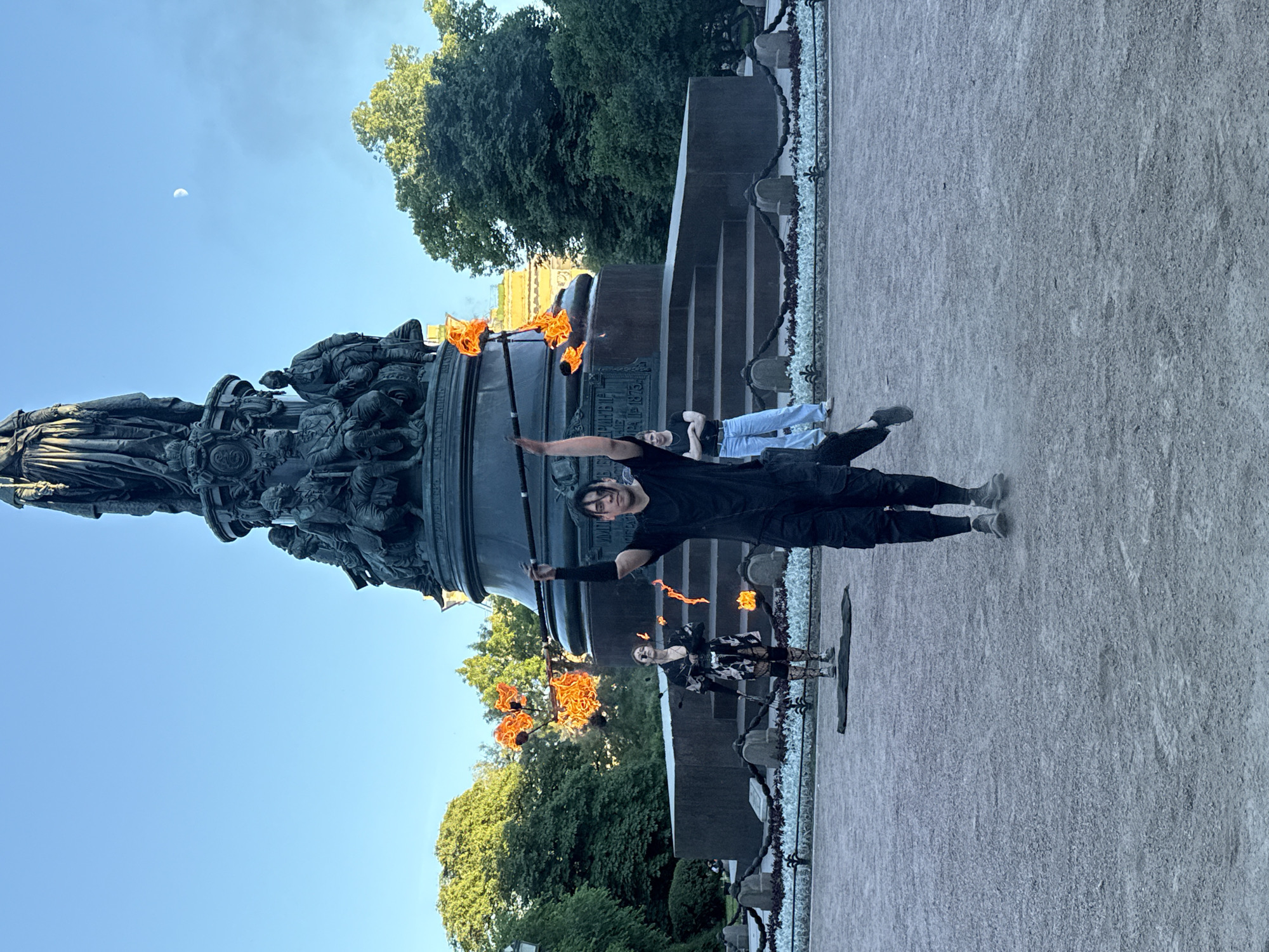 The image shows a group of people performing with fire near a monument in Saint Petersburg, Russia. The monument is a large, dark bronze structure adorned with intricate sculptures of people and animals. It appears to be a historical monument, possibly a memorial. In the foreground, a person in black clothing is holding a flaming prop, appearing to be mid-performance. Further back, two other individuals are also present, one of whom is also engaged in a fire performance with flaming props. One of the performers wears a black and white patterned outfit with fishnet stockings, adding a striking visual element to the scene.
The time of day appears to be late afternoon or early evening, indicated by the warm, golden sunlight casting shadows and illuminating the scene. The sky is a clear, bright blue with a sliver of the moon visible. Lush green trees form a backdrop to the monument, suggesting a park or a public square setting. The ground is paved with small stones or gravel. The overall atmosphere seems energetic and captivating, with the bright flames of the fire props creating a dramatic contrast against the solemnity of the monument and the natural surroundings. There is no visible text in the image.