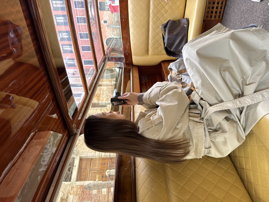 A female individual is seated inside a boat, viewed from the mid-back and right profile. She has long, straight, dark brown hair and wears a light beige trench coat over a grey ribbed sweater. She holds a compact black digital camera with both hands, directed towards the exterior view through the boat's window. The boat's interior features polished dark wood paneling on the ceiling and trim, and quilted yellow upholstery on the seats. Two bags, one grey canvas and one dark brown leather, rest on the seat to her right.
Outside the boat, a scene characteristic of Venice, Italy, is visible. Through the windows, narrow waterways and traditional Venetian buildings are apparent. On the left, a brick building with an arched wooden door and a stone mooring post is partially seen. On the right, buildings with reddish-brown facades and windows with shutters line the canal. The environment is overcast.