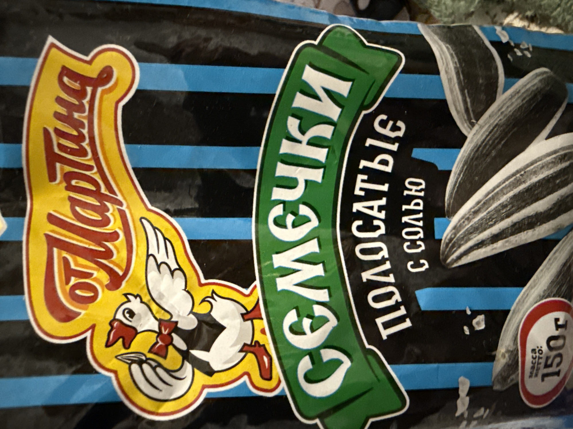 A close-up shot of a bag of sunflower seeds. The bag is black with thin blue vertical stripes. A yellow oval shape at the top of the bag contains red text that reads "Мартина" and a cartoon chicken wearing a bowtie. Below this, a green banner features white text reading "СЕМЕЧКИ". Underneath the green banner, white text reads "ПОЛОСАТЫЕ" and "С СОЛЬЮ" (with salt). Towards the bottom left, a red and white oval label displays "МАССА НЕТТО: 150г". Several sunflower seeds are visible against the striped background.