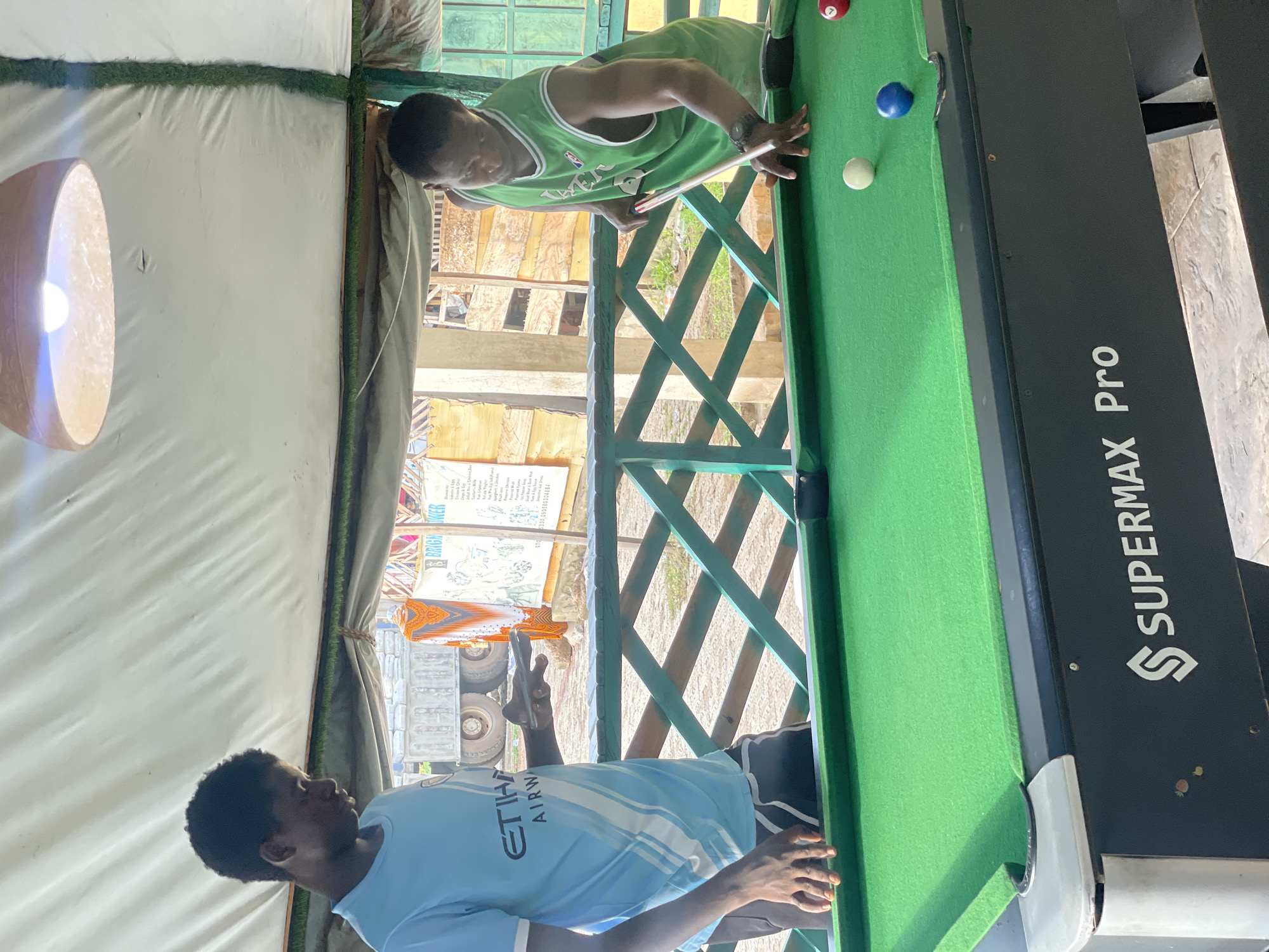 A photograph taken indoors at an elevated level, oriented vertically, depicts two African men playing pool on a Supermax Pro pool table. One man, wearing a green basketball jersey, is positioned at one end of the table, aiming a cue stick at a white ball with a red and blue ball nearby. The other man, wearing a light blue jersey with "Etihad Airways" text, stands at the opposite end, holding a mobile phone. The pool table is green, and the lighting includes an overhead lamp. The background includes green painted wooden framework, and beyond that, a view of the city of Lokoja, Nigeria, including buildings, vehicles, and signage.