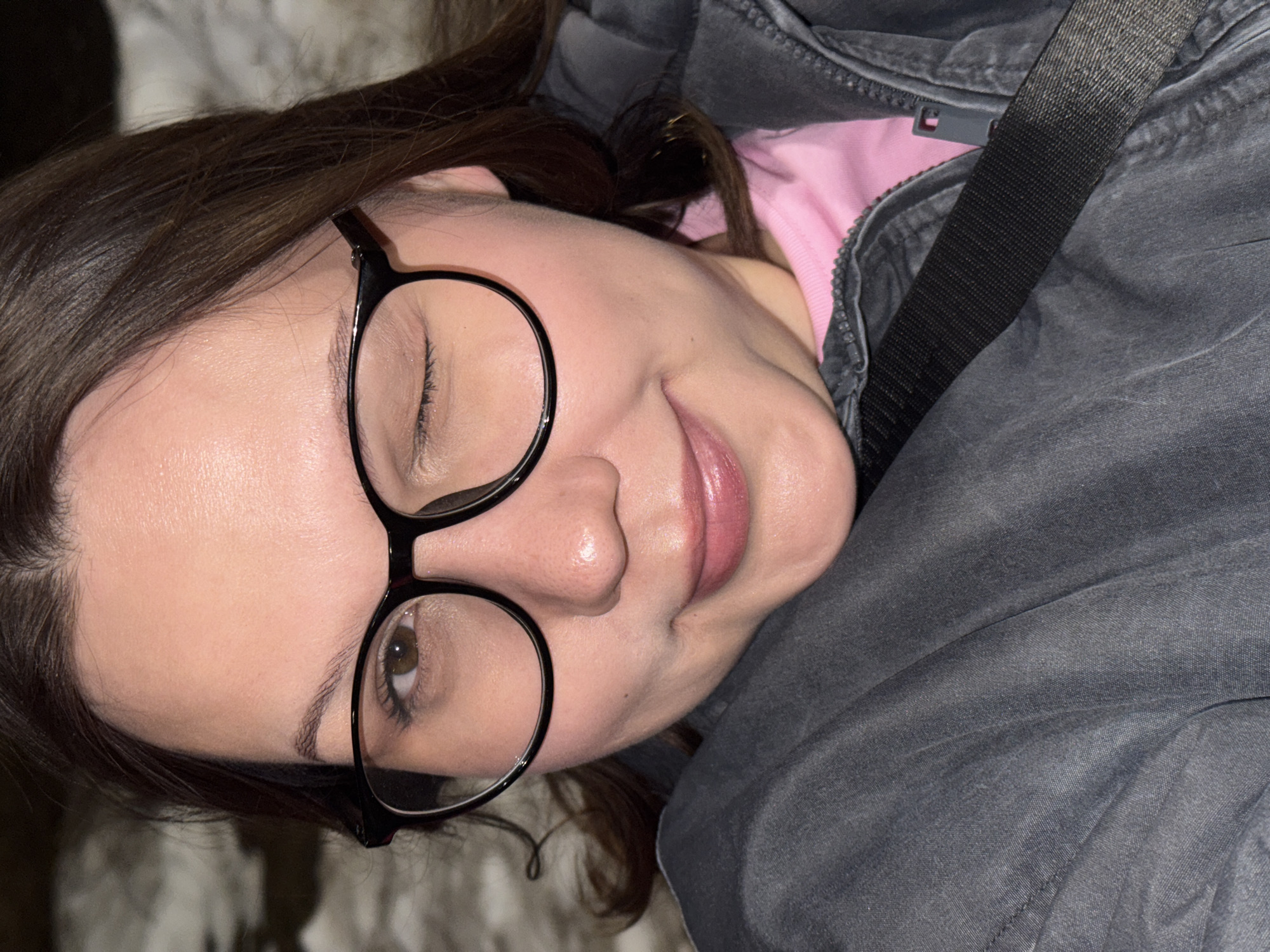 A woman with brown hair and light skin is wearing black-rimmed glasses. She is partially reclined, wearing a pink top underneath a zippered gray jacket. A black strap crosses over the jacket. The background is blurred and indistinct. No information in the image indicates the location is Chaikovsky, Russia.