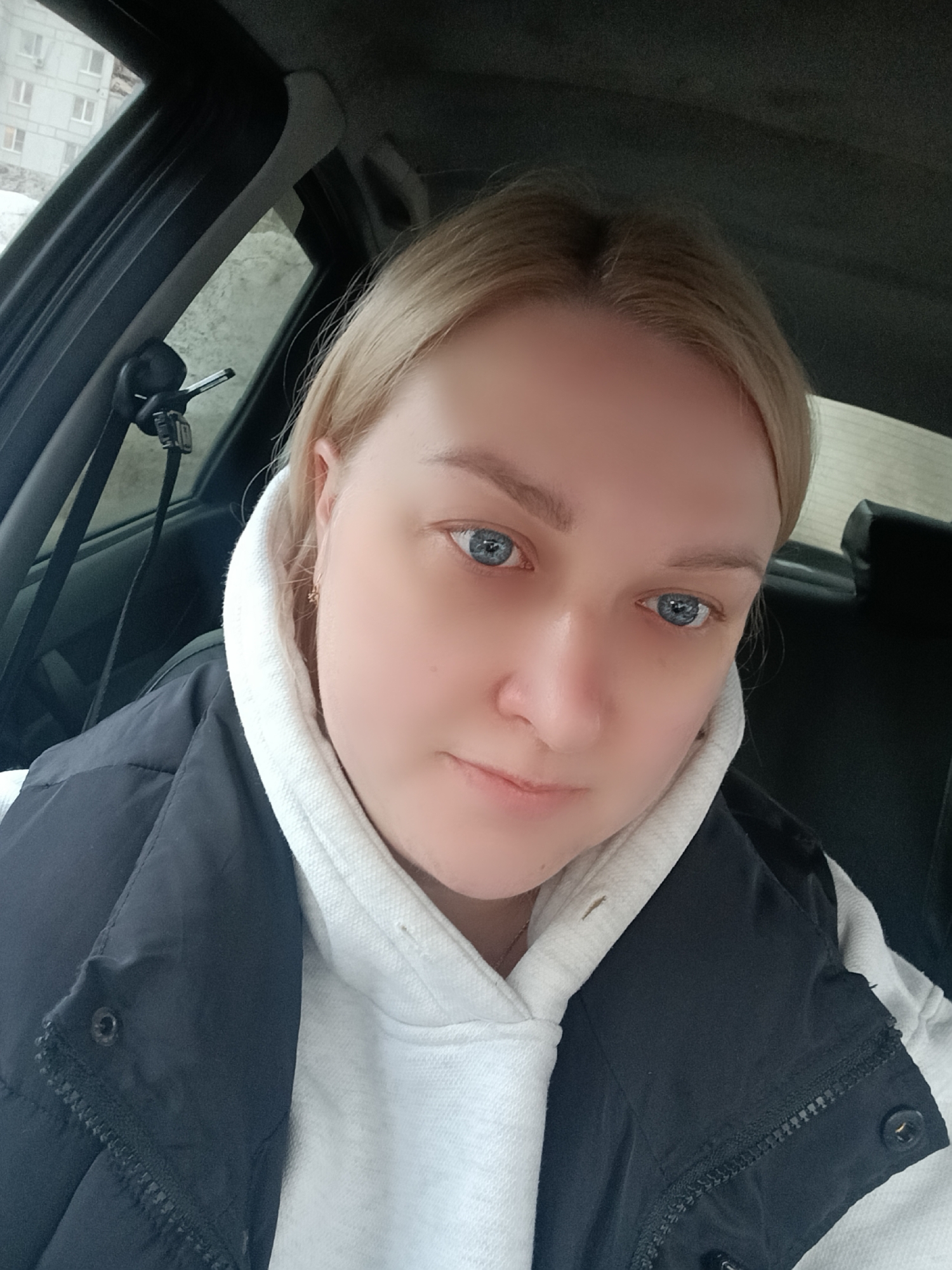 The image depicts an adult female positioned inside a vehicle, directly facing the camera. She has blonde hair, blue eyes, and fair skin, maintaining a neutral expression. She is wearing a white hooded sweatshirt under a dark-colored, possibly black, quilted vest, with a visible zipper and snap button closure. A small, star-shaped gold earring is visible in her right ear, and a thin gold chain is around her neck.
The interior of the vehicle features a dark grey or black headliner and trim. Through the side window on the left, a multi-story light-colored residential building with multiple rectangular windows is visible. The architectural style of this building is consistent with typical urban apartment complexes found in the city of Chaikovsky, Russia. The ground outside the building shows areas of light-colored debris or snow. A black car pillar with a seatbelt mechanism or device mount is visible on the left side of the frame. The overall lighting suggests daylight.