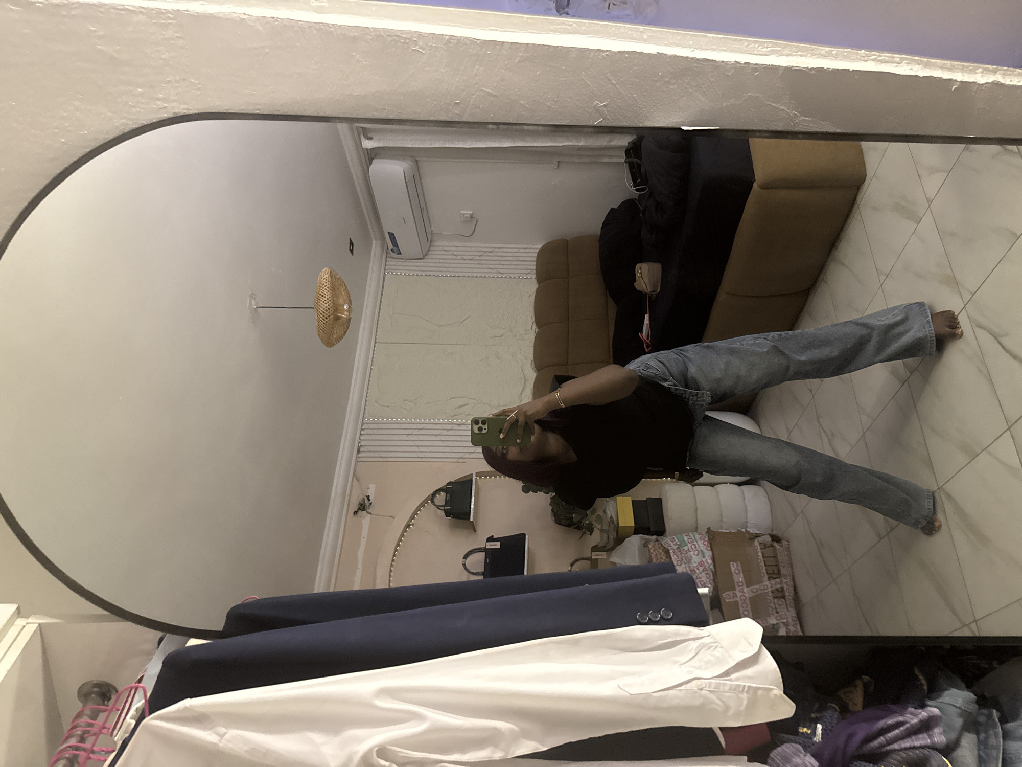 A person is taking a full-body mirror selfie in a bedroom. The person is wearing a black t-shirt and blue jeans. They are holding a green smartphone in their right hand. The bedroom has a bed with brown and black bedding, an air conditioning unit on the wall, and shelves with purses. In the foreground, to the left of the mirror, there is a closet with hanging clothes, including a white shirt and a navy blue jacket. The room has tiled flooring and a wall with textured panels. The mirror itself is large and arched, framed by a white border. The scene appears to be indoors, likely a residential setting. The image does not contain any specific geographical markers to indicate the city of Abuja, Nigeria.