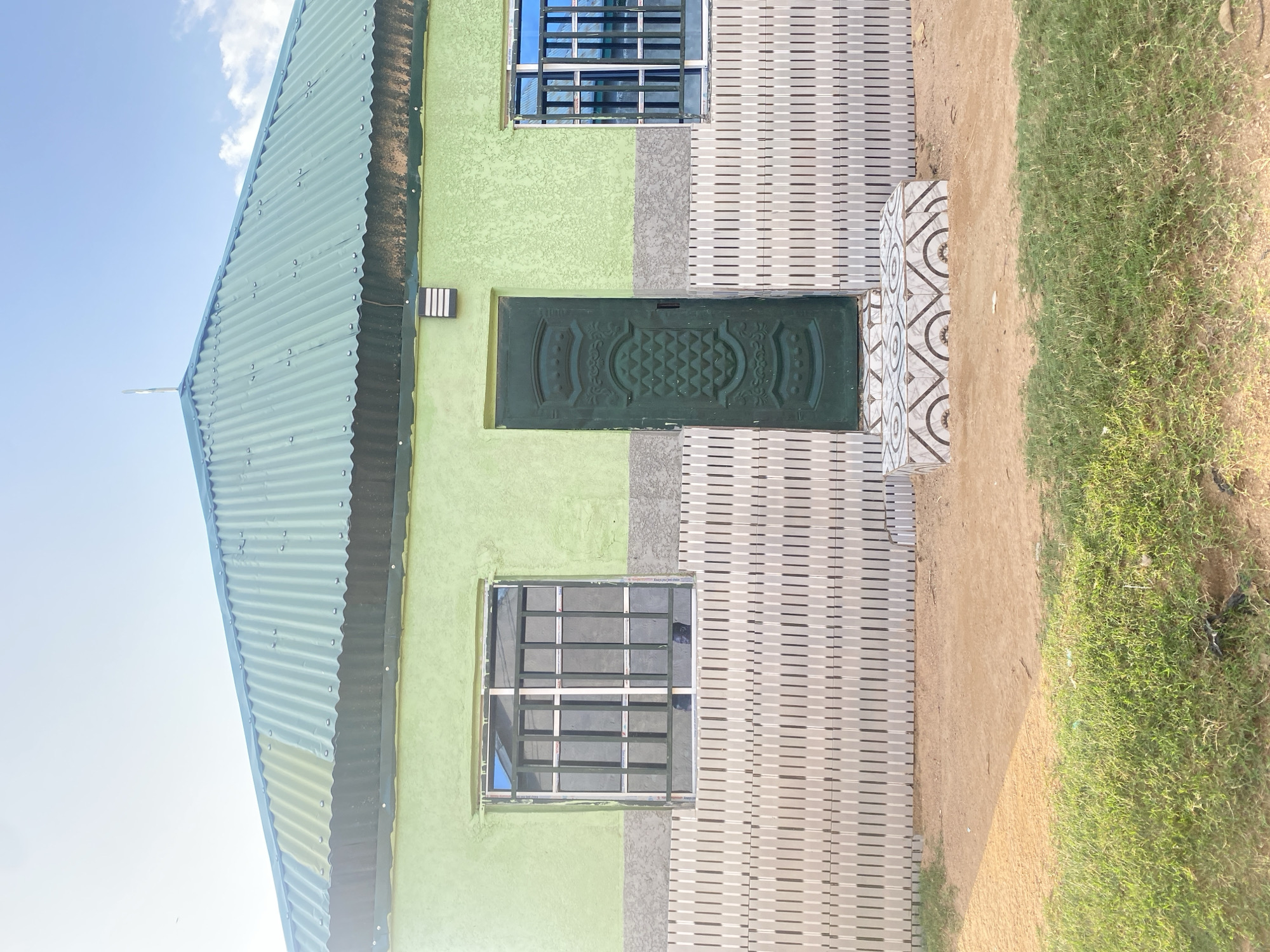 The image depicts a single-story house with a green corrugated metal roof. The exterior walls are painted a light green and a grey-toned textured material is visible below the windows and door. There are two windows with metal security bars and a single dark green door with ornate detailing. In front of the door is a small tiled step. The house is situated on a patch of dry, sandy ground with some green grass in the foreground. The sky above is blue with some scattered clouds. The scene appears to be an exterior shot of a residential building.