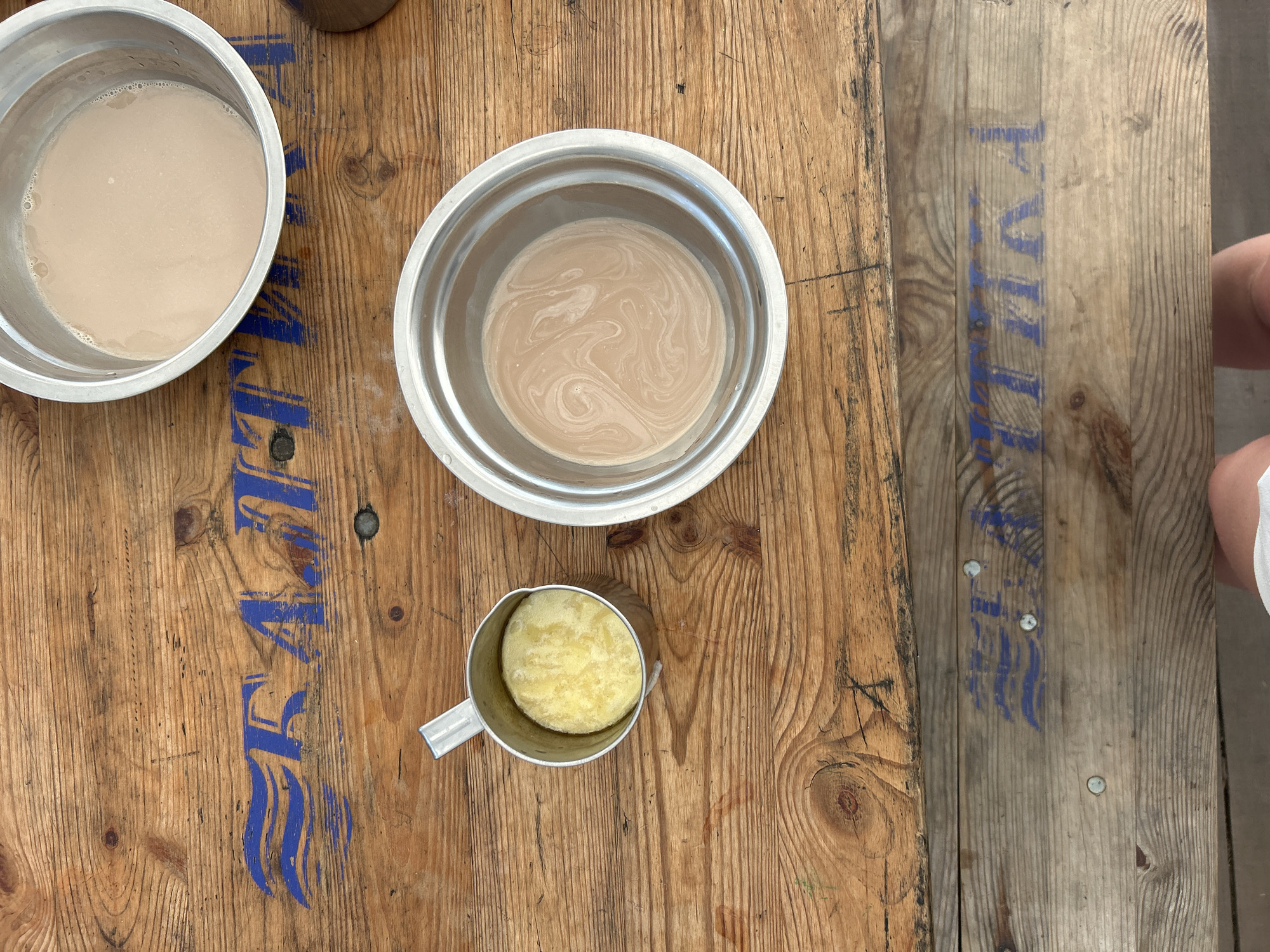 In the city of Nikolo-Petrovka, Russia, on a weathered wooden table, are two metallic bowls containing a light brown liquid, likely a beverage. One bowl is larger and centrally positioned, with swirls indicating it was recently stirred. To its left is a smaller metal cup filled with a solid, yellow substance, possibly butter or a paste. The table itself has visible wood grain and blue stenciled lettering that appears to be a repeating word or logo. In the bottom right corner, a portion of a person's legs wearing white shorts is visible, suggesting someone is seated at the table. The lighting suggests an outdoor or well-lit indoor environment.