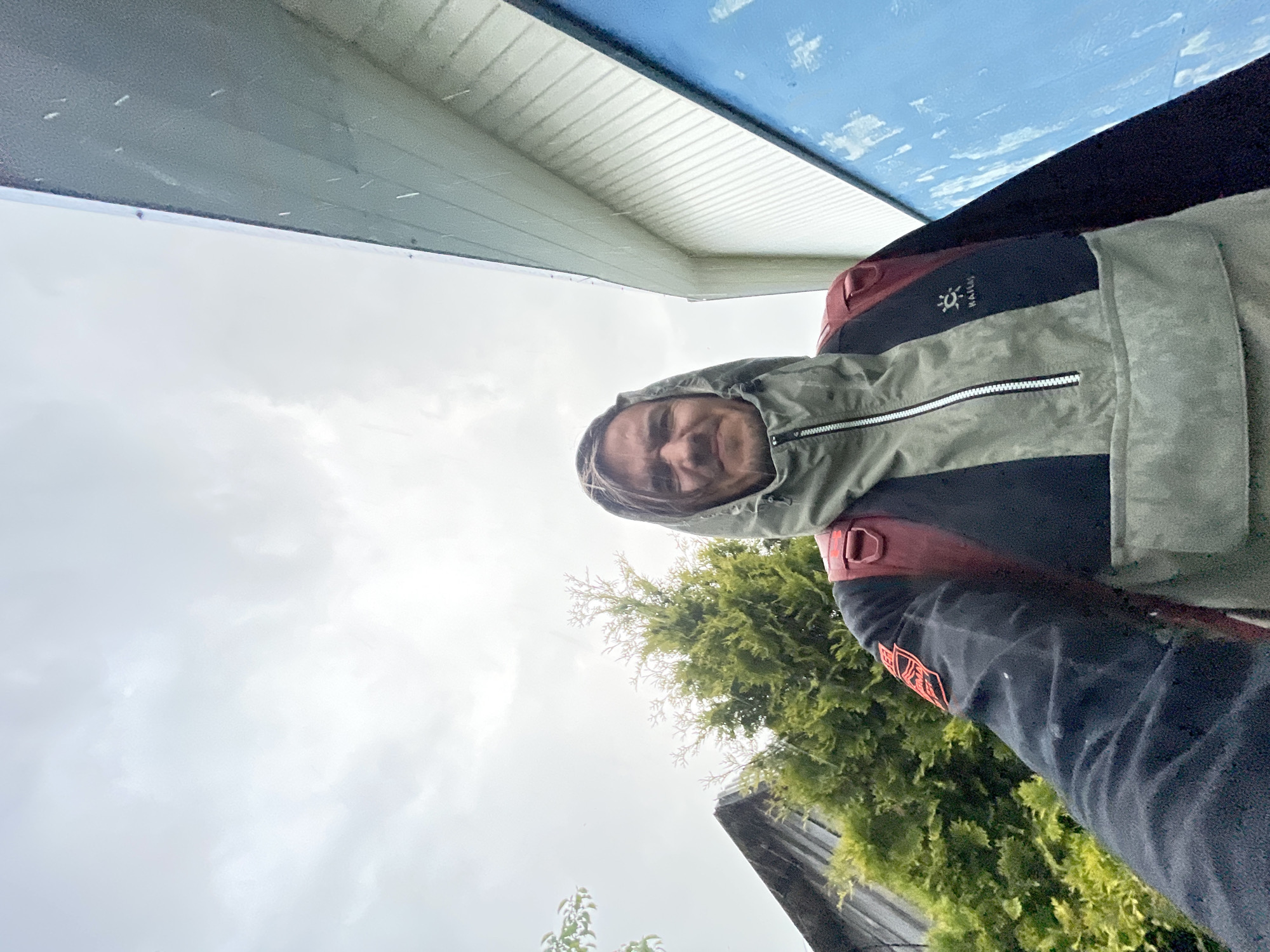 An adult male is depicted in a close-up, upward-angled selfie-style photograph taken outdoors under an overcast sky. The location is in the city of Unknown, Unknown. The man, facing directly forward, has light brown hair falling across his forehead, a light mustache, and a slightly furrowed brow with his mouth gently downturned. He wears a grey-green and black multi-panel anorak-style jacket with a half-zip front and an activated hood. A Haglöfs logo is visible on the jacket's right chest area. A reddish-brown backpack is visible on his shoulders. Small, light-colored specks are visible across the image, consistent with light precipitation.
In the background to the left, a portion of a dark-colored building with a pitched roof is visible, along with dense green coniferous trees. To the right, a light-colored building with horizontal siding is partially visible, adjacent to a large, vertical blue surface displaying scattered white markings. The sky above is uniformly light grey and cloudy.
