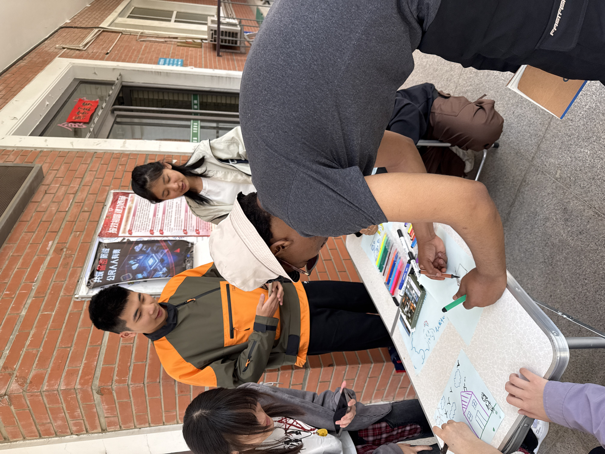The image captures a group of five young people engaged in an outdoor drawing activity next to a brick building, likely on a campus or public space in Daxing District, China.
At the focal point, a person wearing a dark grey t-shirt and a light cap is leaning over a white foldable table, drawing on a piece of paper with a green marker. A collection of colorful markers and a marker case are spread across the table. Multiple hands are visible on the table, indicating collaborative drawing on several sheets of paper; one drawing depicts a house.
To the left of the table, a man in an orange and dark green jacket observes the activity with a slight smile. Next to him, a woman in a grey jacket and plaid shirt looks down at a smartphone. In the background, another woman in a white jacket smiles as she looks towards the drawing activity. Further back, a brown mannequin head or similar object is placed on a chair.
The setting includes a building with a red brick facade and windows. A red decorative item with Chinese characters hangs inside one window. On the brick wall, two posters are visible: one with the text "投效祖国 青春无悔" (Serve the motherland, youth without regret), and another stating "共筑品质课堂 公民人人有责" (Jointly build quality classrooms, every citizen has a responsibility). The ground is paved with light-colored tiles. The scene is brightly lit, suggesting daytime. The individuals appear focused and engaged in the creative activity.