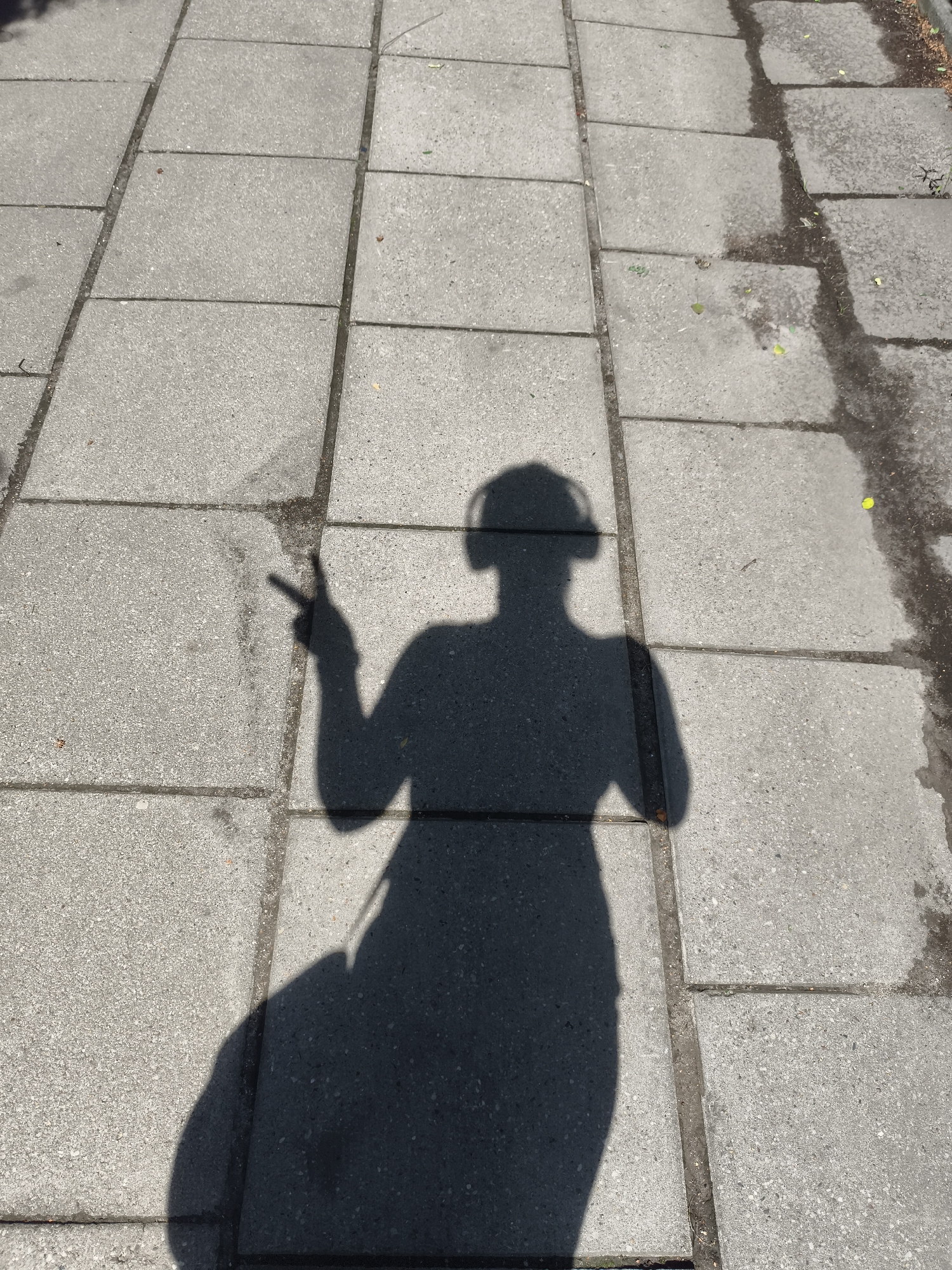 The image is a high-angle shot of a shadow of a person on a paved walkway. The walkway is made of rectangular concrete slabs laid in a grid pattern. There are some dark, wet patches on the walkway, suggesting recent rain or water spillage. The shadow depicts a person wearing headphones, with their right arm raised and two fingers extended in a peace sign. The person is also carrying a bag on their left shoulder, indicated by the shadow of the strap. The background appears to be a city environment, possibly in Gdańsk, Poland, given the context. The lighting suggests it is daytime, with the sun casting a distinct shadow.