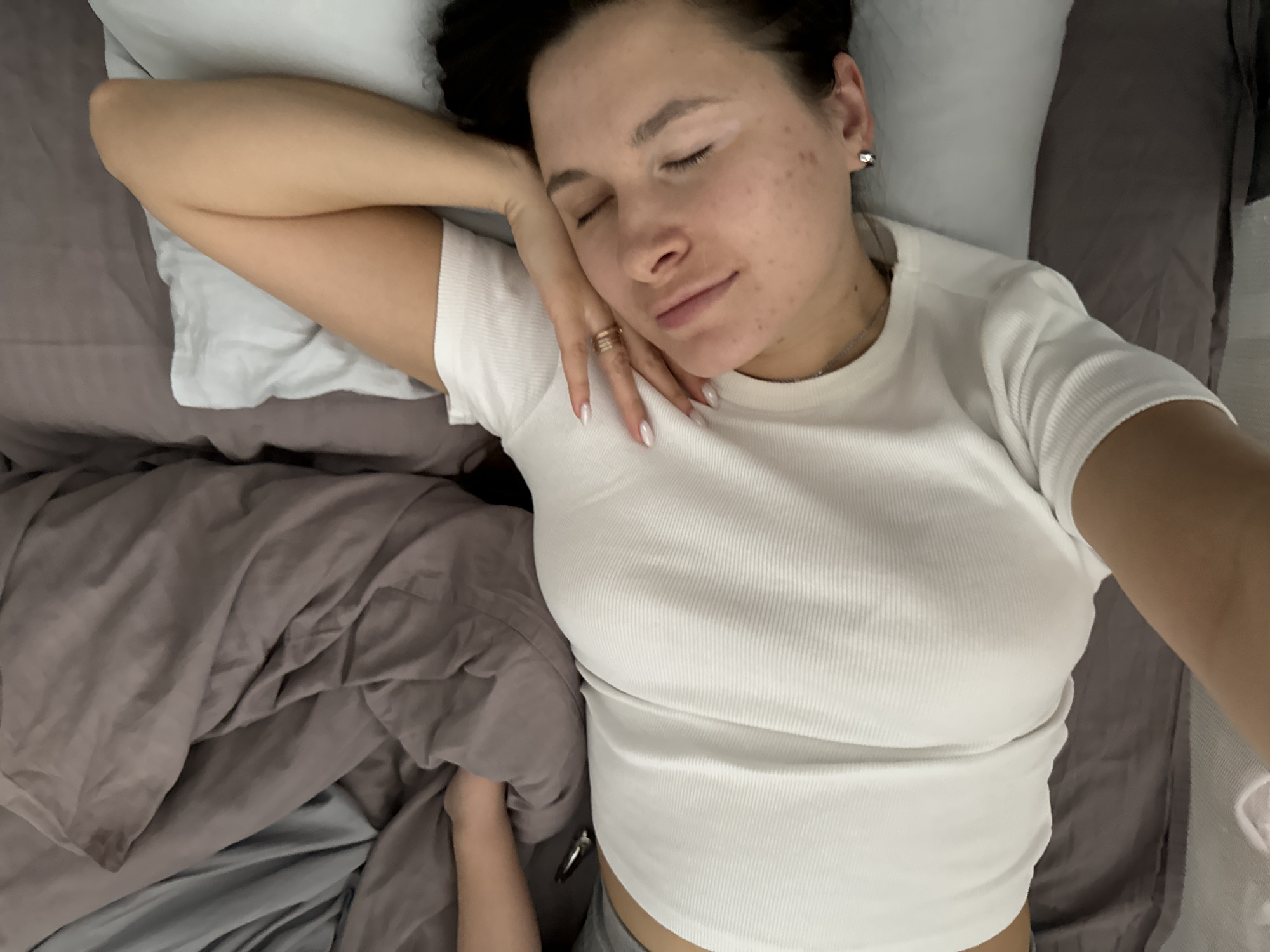 A young woman is lying on her back on a bed. Her eyes are closed, and she appears to be sleeping. She is wearing a white t-shirt. Her left hand is resting on her face. She has several rings on her fingers. She has dark hair and fair skin. The bed has a gray sheet and a white pillow. There is a brown blanket rumpled on the left side of the frame. A small child's arm is visible on the left side of the image, under the blanket. The location is possibly Akulovo, a city in Russia.