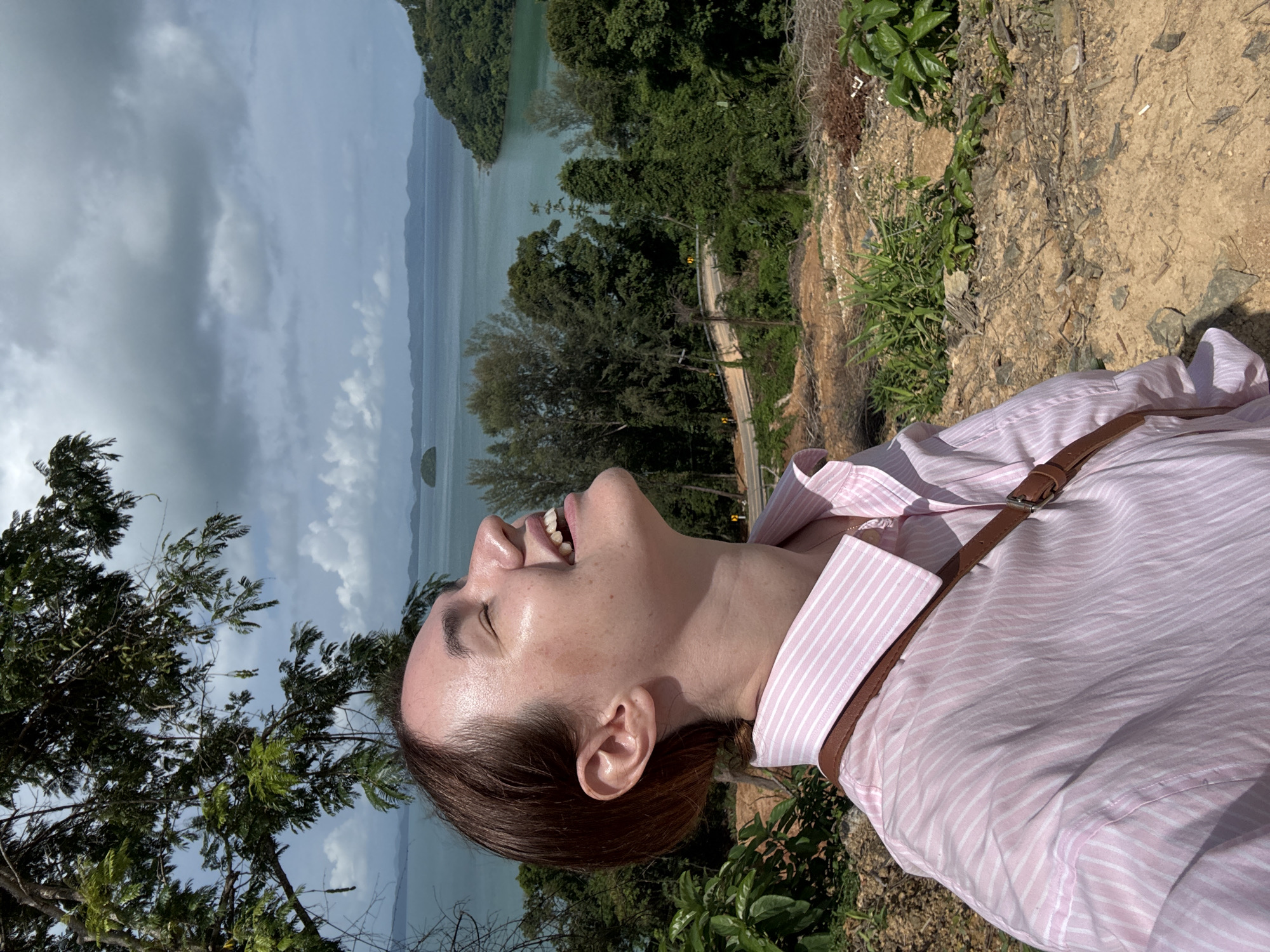 A young woman is captured in a close-up, side-profile shot, looking upwards with her eyes closed and a joyful expression. She has light skin, short brown hair, and is wearing a pink and white striped button-up shirt. A brown strap from a bag crosses her chest. The background depicts a lush, green landscape with dense trees and a winding road. Beyond the trees, a body of turquoise water is visible, dotted with islands and distant mountains under a cloudy sky. The setting is likely an elevated viewpoint in Pa Khlok, Thailand, offering a scenic coastal vista. The overall impression is one of peaceful enjoyment and connection with nature.