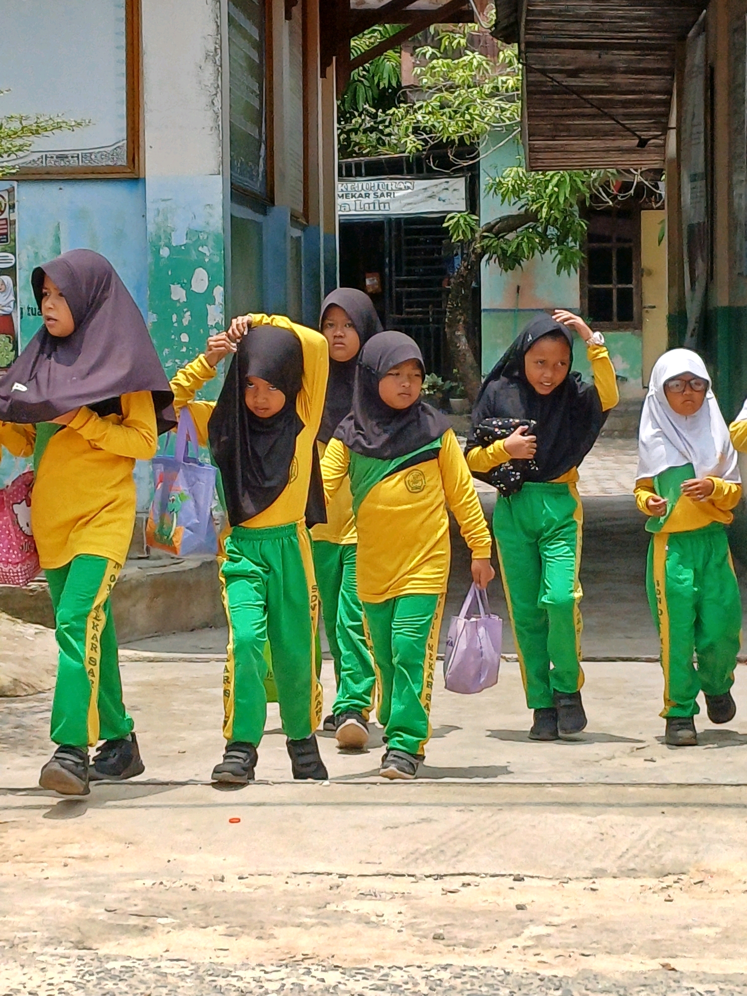 Six young girls wearing yellow long-sleeved shirts with green trim and green pants with yellow trim walk along a paved path. All are wearing headscarves, with four wearing black and one wearing white. The girl on the far right also wears glasses. The girls appear to be in a school setting, as indicated by a building with a blue and green painted facade and a wooden awning in the background. The environment suggests an outdoor, possibly tropical, location. One girl is carrying a light purple bag, and another has a pink bag with a cartoon character on it. The ground is a light gray, textured concrete. The overall scene depicts children walking, likely to or from school.