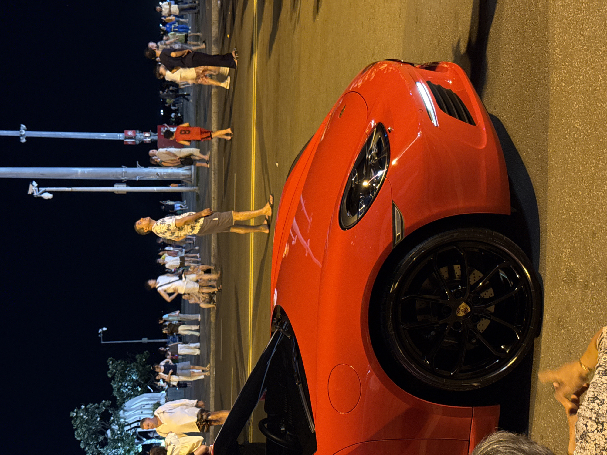 This image shows a red sports car, possibly a Porsche, parked on a street at night. The street is lined with people, some of whom are casually standing and talking, while others appear to be walking. The background suggests an urban environment with tall poles that could be streetlights or lampposts. The people in the image are dressed in casual summer attire, indicating a warm climate. The car is the main focus in the foreground, with its sleek design and bright red color standing out against the darker background. There is a crowd of people further down the street, suggesting a lively atmosphere, possibly a night market or a gathering place. The overall impression is that of a vibrant city scene at night.
