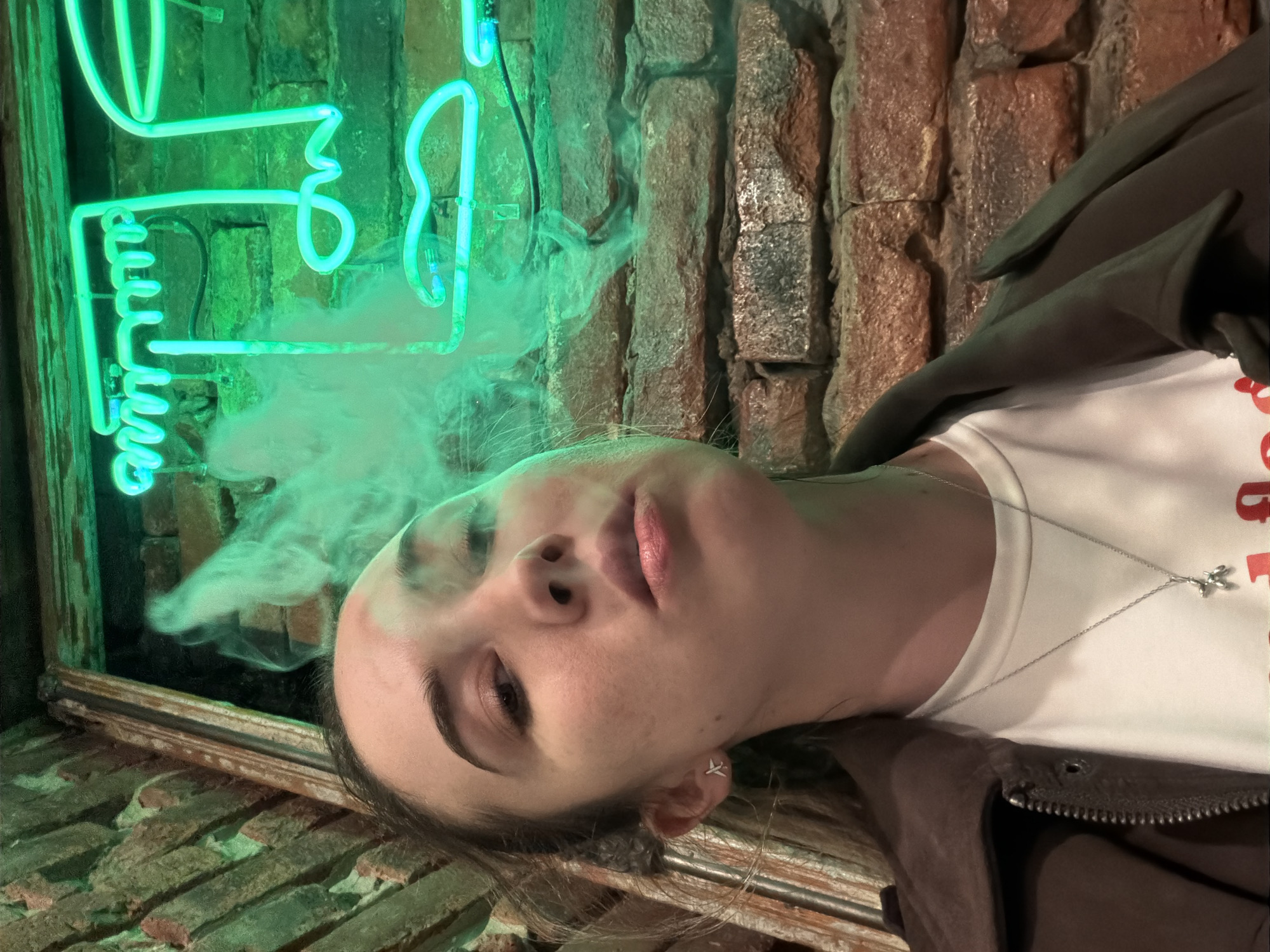 A woman is seen lying on a brick surface, exhaling smoke. She wears a white t-shirt and a brown jacket. A silver necklace with a pendant is visible around her neck. A neon sign with a stylized face is on the wall behind her. The location appears to be indoors, possibly a bar or lounge, with exposed brick walls. The location is Rostov-on-Don, Russia.