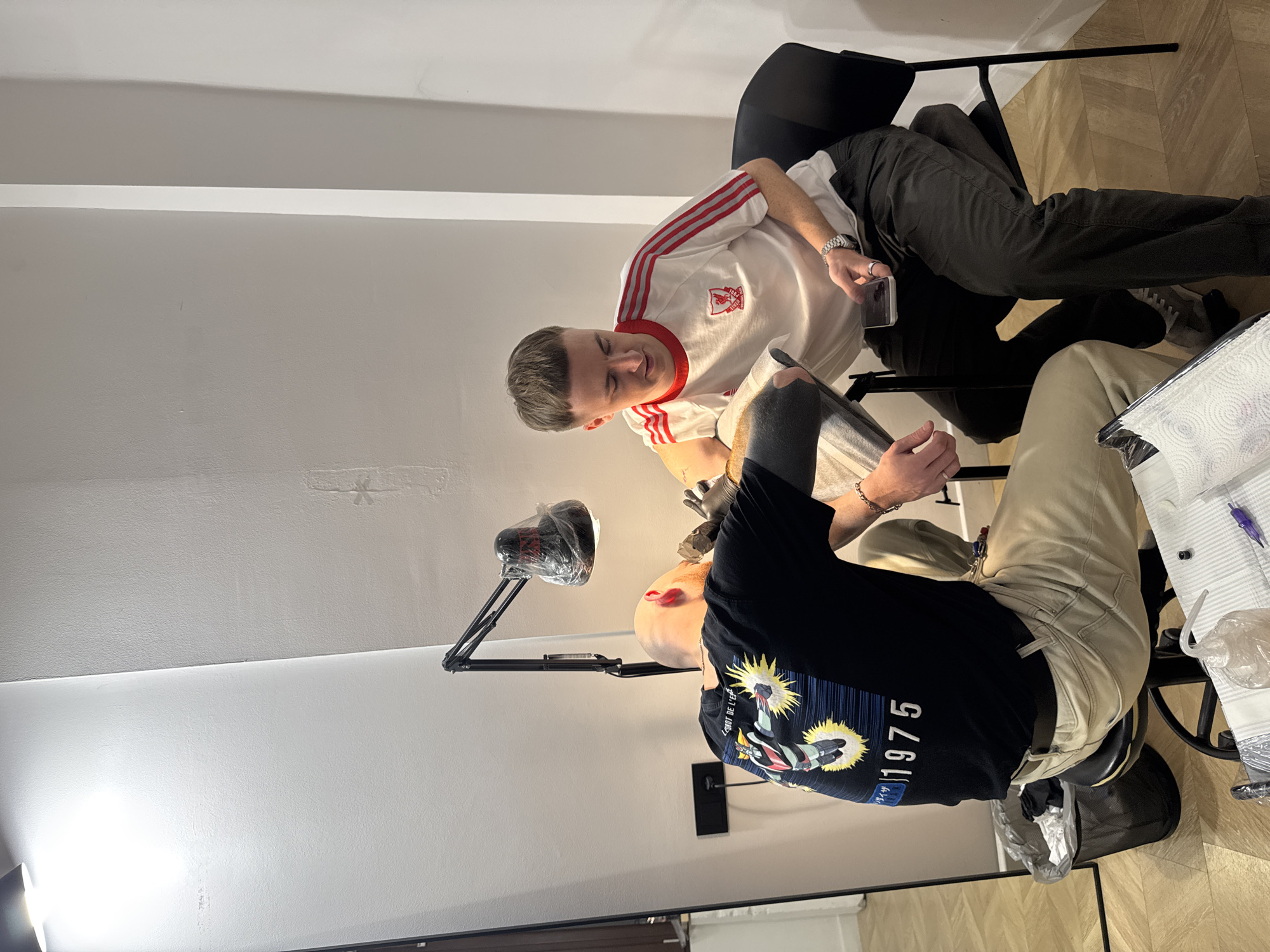 Two men are in what appears to be a tattoo studio. One man wearing a white shirt with red stripes and dark pants is sitting in a black chair and looking down at the other man, who is wearing a black shirt with a graphic and khaki pants and is seated in a tattoo chair. The tattoo artist has black gloves on, and there is a lamp positioned above them. Tattooing supplies and paper towels are on a stand next to the tattoo artist. The location is likely in Tbilisi, Georgia. The floor is light-colored wood. The walls are light grey, and the setting appears to be indoors. The man in the white shirt is holding a smartphone.