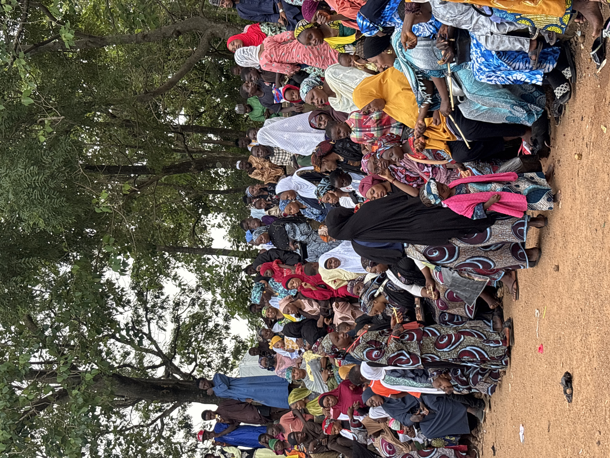 A large group of people, primarily women and children, are gathered outdoors, seated on a tiered, earth-toned embankment. They are dressed in colorful, patterned traditional clothing, with many women wearing headscarves. The environment is rural, with lush green trees providing a backdrop and canopy overhead. The ground in the foreground is bare earth, with some scattered debris. The people appear to be observing something or participating in an event, with some looking towards the left of the frame. The overall impression is one of a community gathering. The location is the city of Jos, Nigeria.