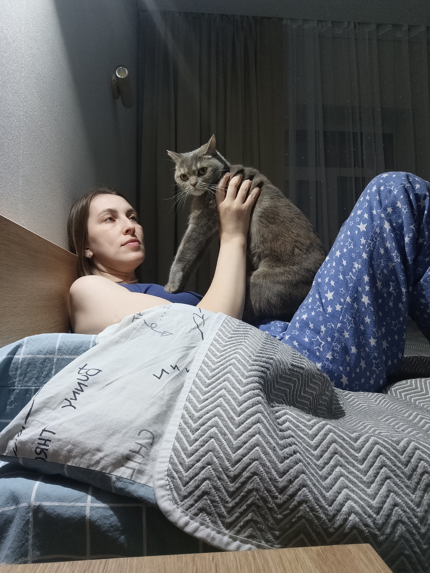 The scene depicts an adult female and a grey shorthair cat on a bed within a residence in Chaikovsky, Russia. The female, appearing in her 20s or 30s, lies on her left side, propped against a light wooden headboard, wearing a dark blue top and blue pajama bottoms with white star patterns. Her right hand rests on the cat, which stands on her lap/torso, facing forward with its head slightly tilted and yellowish-green eyes. The bedding includes a blue and white checkered sheet, a white pillow with black Cyrillic text, and a grey chevron-patterned blanket covering the lower body. The background features a textured grey wall, a wall-mounted sconce with a yellow-toned bulb, and dark curtains covering a window, indicating a low-light indoor environment.