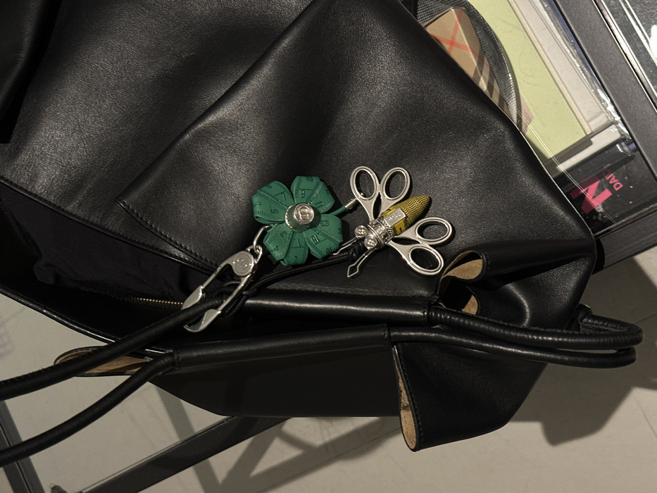 The image displays a black leather handbag resting on a reflective surface. A silver safety-pin style clasp is affixed to one of the bag's handles, from which two decorative charms are suspended. The upper charm is a four-leaf clover shape, constructed from green measuring tape with visible numerical markings, centered by a silver button embossed with the "CD" logo. Below this, a second charm depicts a stylized insect with silver-colored wings resembling small scissors and a textured yellow body, also fashioned from measuring tape. In the background, a partial view reveals a cardboard box featuring the distinctive Burberry Nova Check pattern. Adjacent to this, a dark object with partially visible pink text, "DAI N", is discernible through a transparent material, consistent with a display case. The scene is set within a luxury retail environment, likely a high-end department store or boutique in a prominent shopping district of London, United Kingdom, such such as Knightsbridge (Harrods) or Oxford Street (Selfridges).