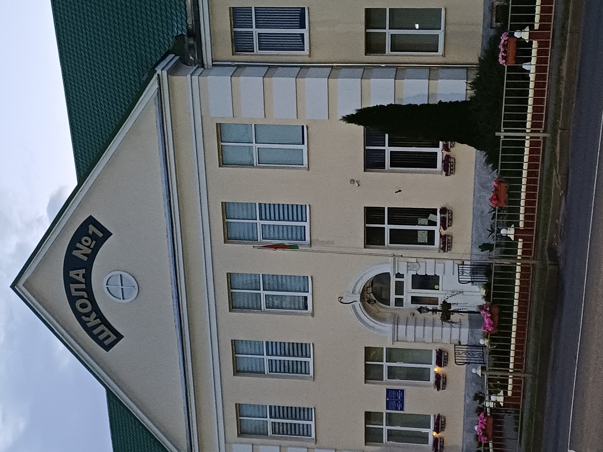 The image shows the facade of a school building, identified by the Cyrillic lettering "ШКОЛА № 1" (School No. 1) above the entrance. The building is light yellow with a green tiled roof and white trim around the windows and corners. There are multiple rows of windows on the facade, some with blinds. A flag is visible on the right side of the upper floor. In front of the building, there is a fence with planters containing pink flowers, and a pathway leads to the entrance. A tall, dark green evergreen tree stands to the right of the entrance. The building appears to be located in Kobryn, Belarus, as indicated by the school's name and numbering. The sky is overcast, suggesting a cloudy day.